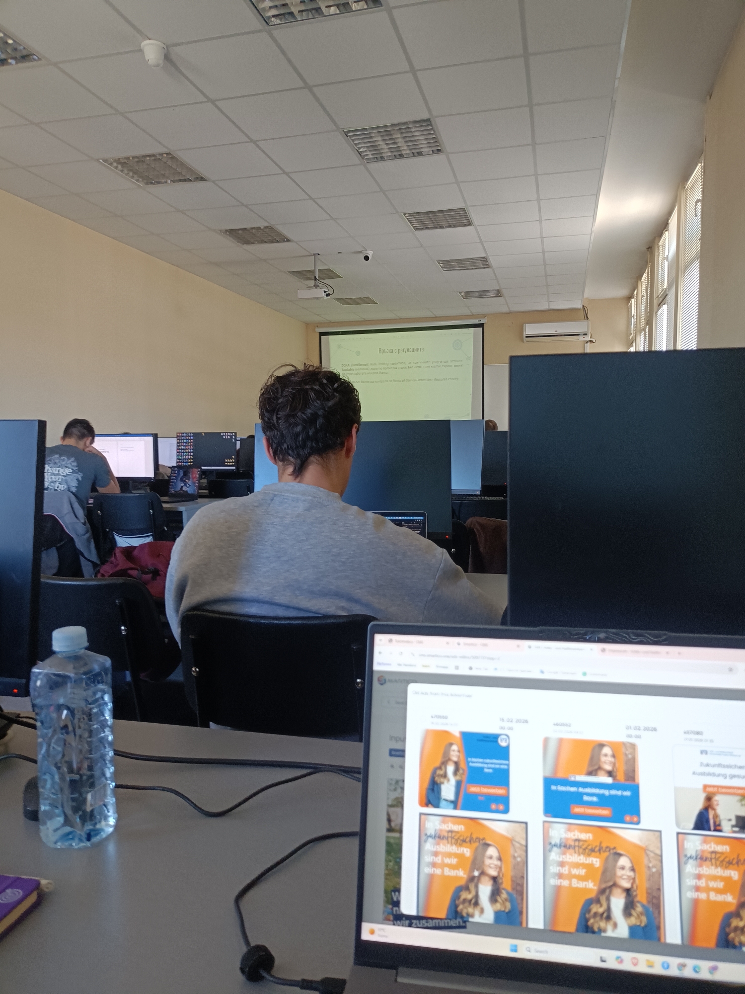 The photo shows the interior of a computer lab, likely a classroom or office, in Unknown, Unknown. In the foreground, a laptop displays a webpage filled with recruitment advertisements, featuring a woman. Next to the laptop, a water bottle and other cables are present on the desk. A man with curly hair is seated in front of a computer monitor, facing towards a projector screen displaying text in a foreign language. Another man can be seen in the background, also seated at a computer. The room is lit by overhead fluorescent lights, and natural light streams in through windows on the right side of the image. The overall impression is of a learning or working environment with technology.