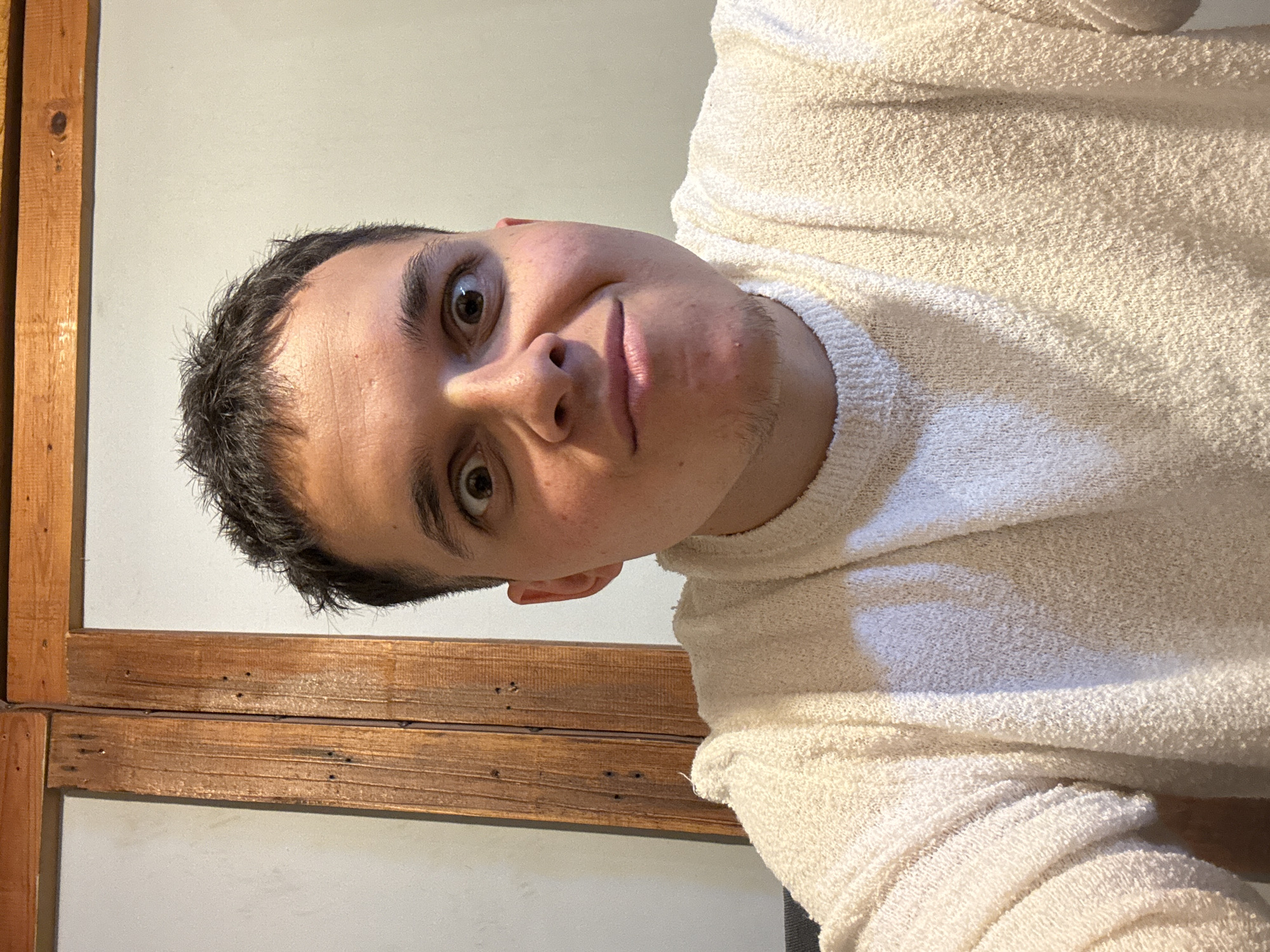 A person wearing a white sweater is looking directly at the camera. The person is indoors, with a wooden structure and a white wall behind them. The photograph does not contain any information to indicate that the location is Kostinbrod, Bulgaria.
