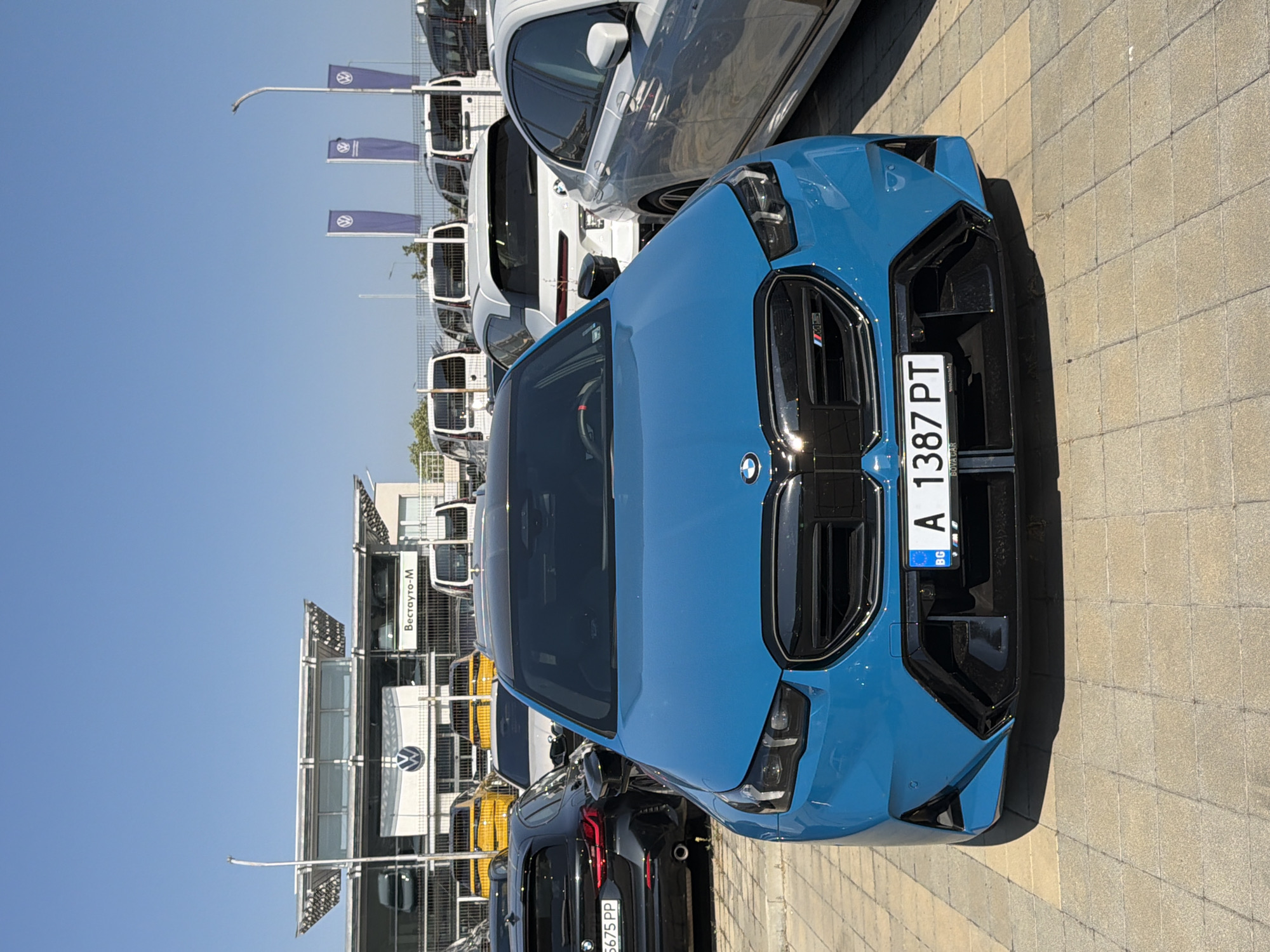 A bright blue BMW M electric vehicle is parked in front of a car dealership under a clear blue sky. The dealership has a modern architectural design with a large glass facade and a prominent "Volkswagen" logo displayed. Several other vehicles, including yellow cars and white vans, are visible in the background. The BMW has a license plate that reads "BG A 1387 PT". The ground is paved with square concrete tiles. The dealership's name, "Вестауто-М", is visible on a sign. The scene appears to be a sunny day in Burgas, Bulgaria, likely at a car lot.