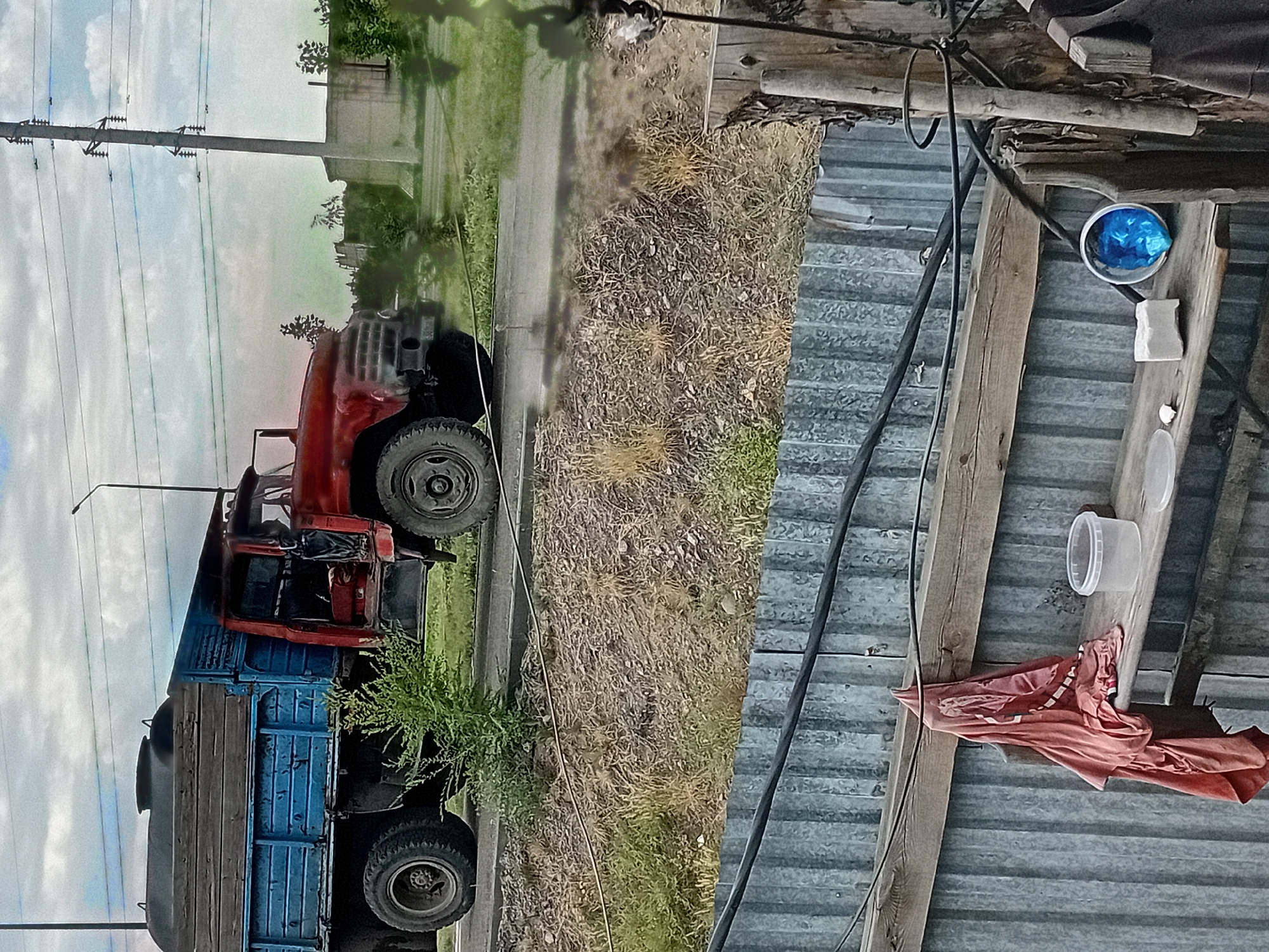 The image shows a side view of a red truck with a blue flatbed carrying a large wooden tank. The truck is parked on a paved surface next to a grassy, dry embankment. In the background, there is a utility pole with power lines, some sparse vegetation, and a building. The foreground shows a corrugated metal fence and a wooden shelf with several items on it, including a pink garment hanging from a hook, a clear plastic container, a white object, and a blue object inside a white container. The sky is cloudy. The scene is set in the city of Semey, Kazakhstan, likely in an industrial or semi-rural area given the presence of the truck and utility infrastructure. There are no people visible in the image.
