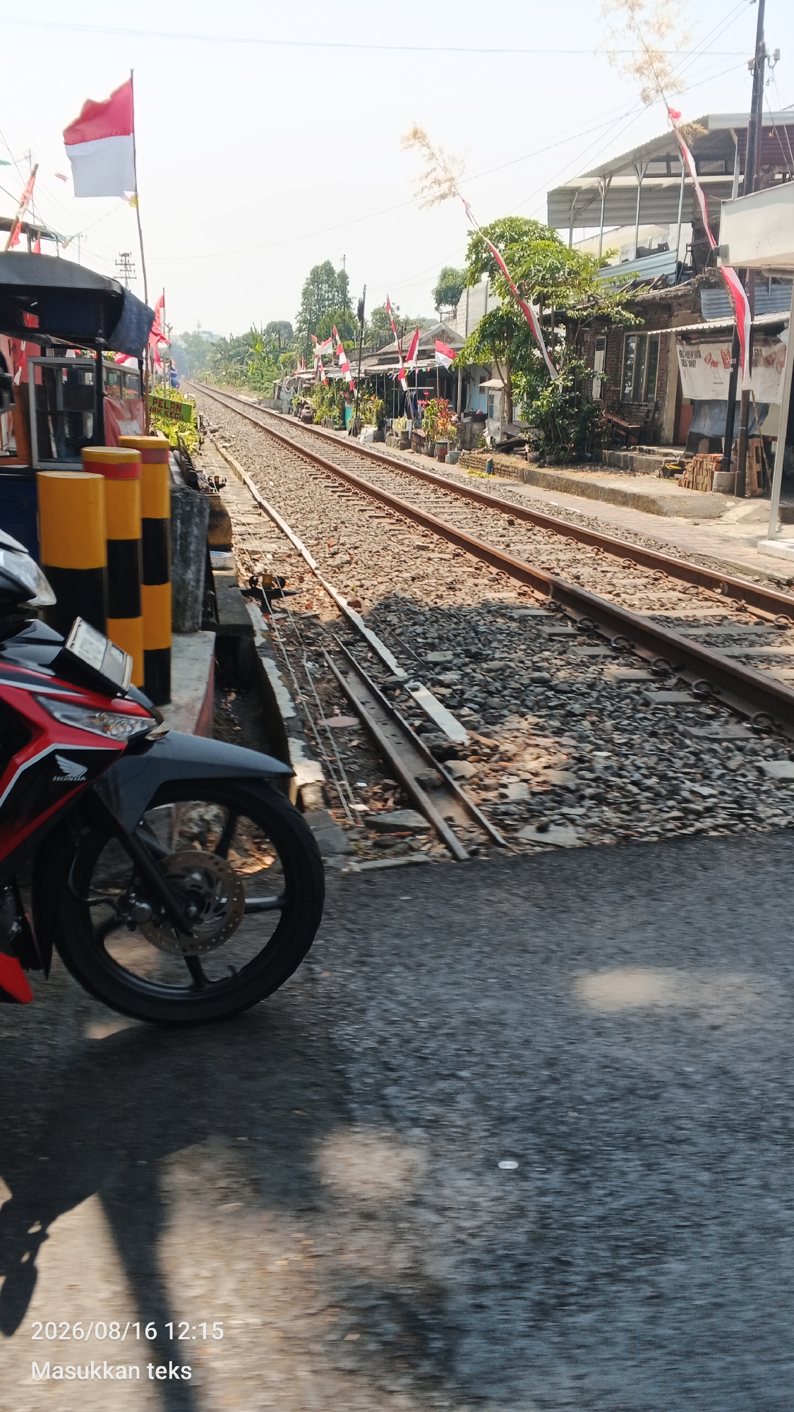 A low-angle, eye-level, outdoor, daylight shot captures a railway track stretching into the distance, lined with residential buildings adorned with Indonesian flags. The flags, red and white, are tied to poles and strung across the street, indicating a national celebration. The tracks are a mix of ballast and gravel, with some overgrown weeds visible. To the left, a motorcycle's front wheel and part of its red and black fairing are in the foreground. Behind the motorcycle are yellow and black striped bollards and a blue structure, possibly part of a railway crossing gate. The background features a hazy sky and lush green trees. The timestamp "2026/08/16 12:15" and text "Masukkan teks" are visible in the bottom left corner. The scene appears to be on a street in the city of Malang, Indonesia.