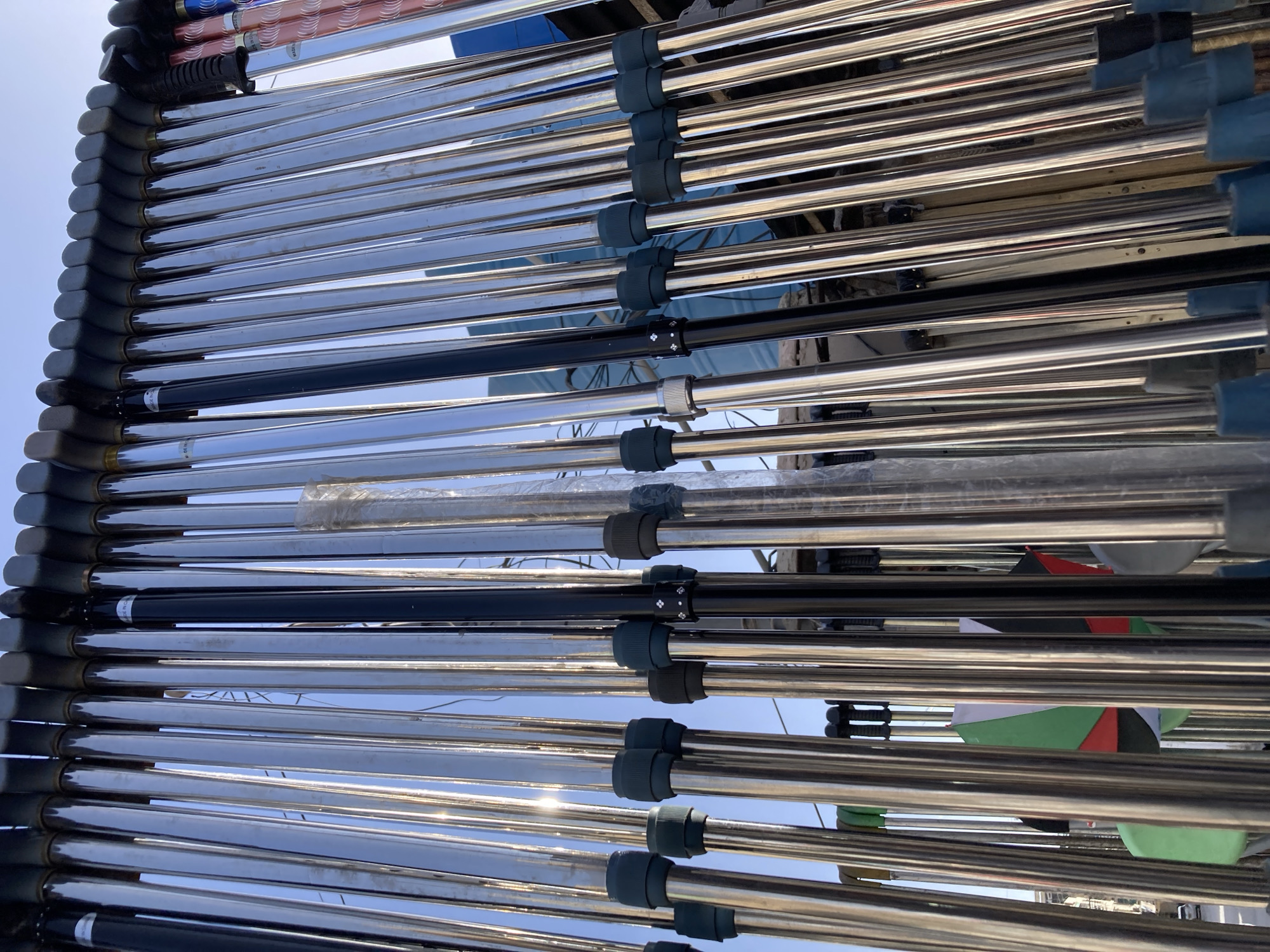 A high-angle, close-up shot depicts a collection of parallel, polished metal tubes, possibly part of a solar water heating system, secured within a dark gray frame with black or dark gray connection joints. The tubes reflect the sky, which appears light blue. Some tubes are darker in color, either black or dark blue. Debris, potentially building materials, is visible between the tubes. Reflections hint at an urban environment. The location cannot be determined from the image.