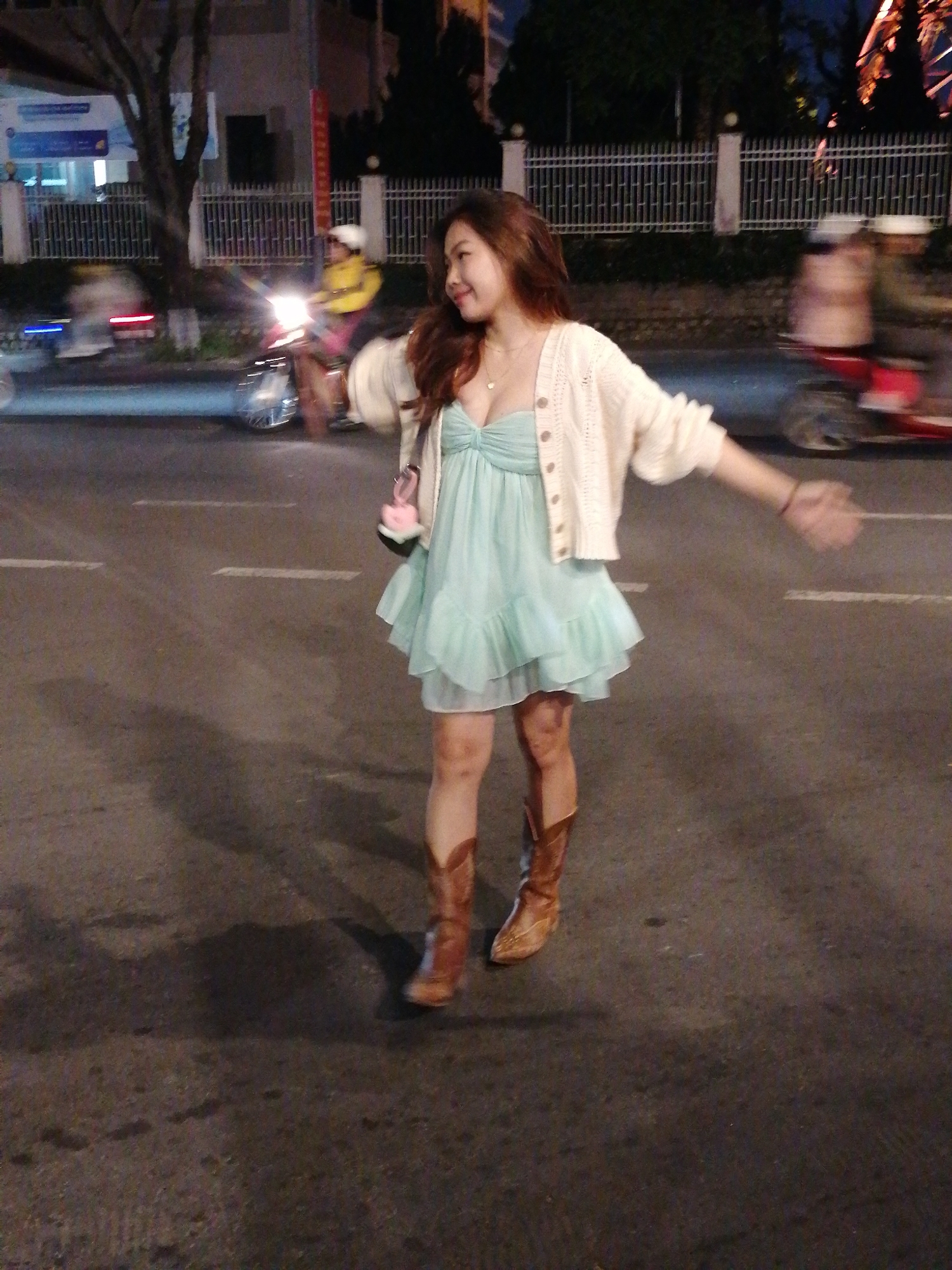 A woman with long, wavy brown hair is standing on a street at night, looking to her right with a slight smile. She is wearing a light blue, ruched, strapless dress with a tiered, ruffled skirt, and a cream-colored, knit cardigan. Her arms are outstretched to her sides, and she has a small pink purse hanging from her left shoulder. She is wearing brown cowboy boots. The street is paved, with white lines marking lanes. In the background, a white metal fence borders the street. To the left, there are blurred lights from vehicles, including a motorcycle with its headlight on. To the right, there are more blurred vehicles. The overall scene suggests an urban setting at night, possibly a street in Da Lat, Vietnam, given the typical attire and traffic observed in such locations. The lighting is dim, typical of nighttime city environments.