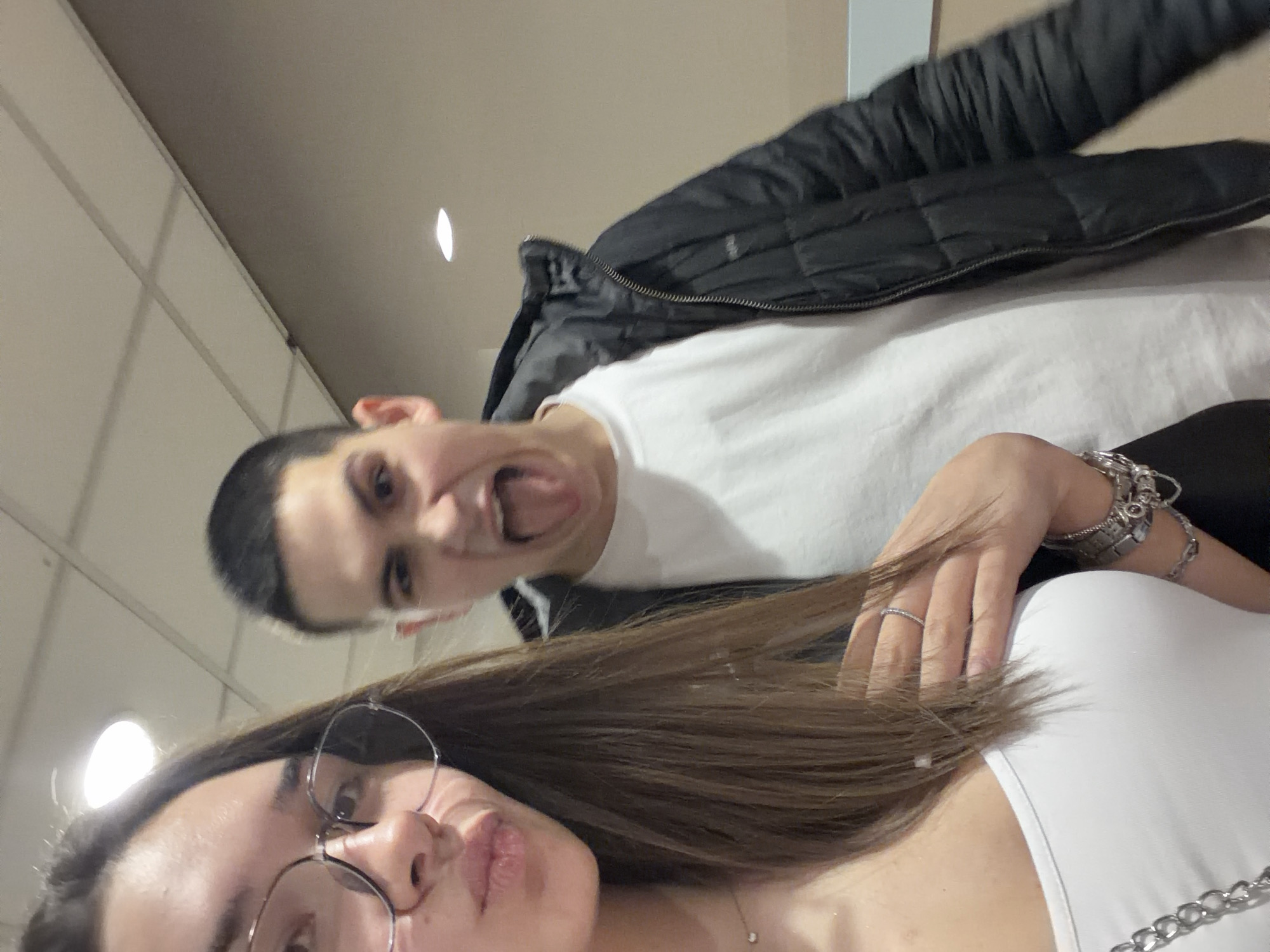 A close-up, selfie-style photograph depicts two individuals indoors. The foreground subject, a female, is positioned on the left, displaying long brown hair, round eyeglasses, a white top, a thin necklace, and multiple bracelets and a ring on her left hand. Her mouth is slightly pursed. Behind and to the right, a male subject with short dark hair is partially visible, wearing a white t-shirt under an open black padded jacket. This male is making a playful expression, sticking out his tongue towards the camera. The background consists of plain, light-colored walls and a dropped ceiling featuring grid panels and recessed circular light fixtures. No specific identifying markers are present within the image to pinpoint a location within the city of Sofia, Bulgaria.