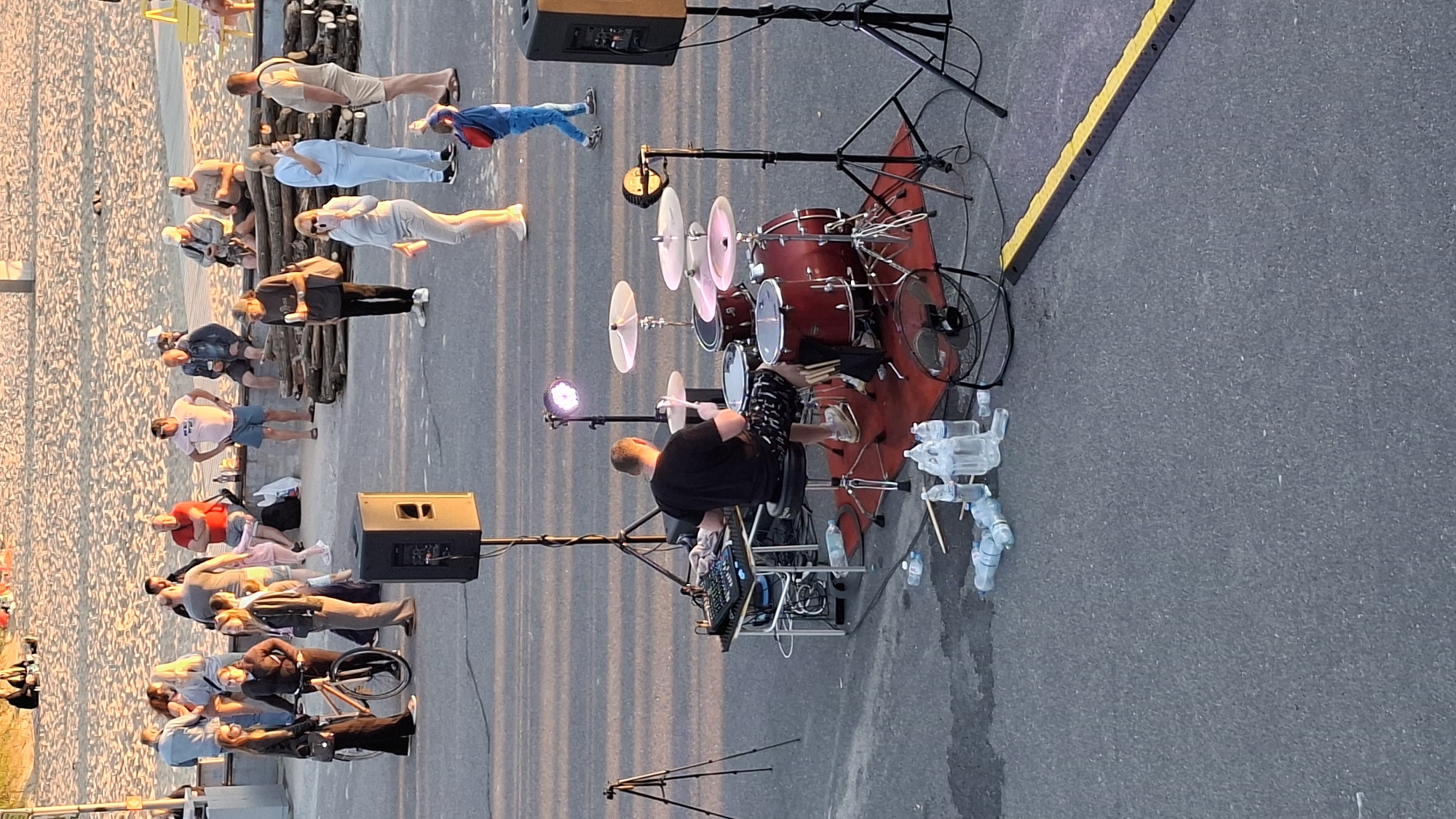 This image shows a street scene in Tallinn, Estonia, with a sandy beach in the background. In the foreground, a person is playing a drum set on a red rug. The drum set is red and has several cymbals. There are two large speakers on stands, a mixing board, and a fan. Several bottles of water are scattered on the ground. In the middle ground, a group of people are standing and sitting on a bench. One person is pushing a bicycle. In the background, more people are seen on the beach. The lighting suggests it is late afternoon or early evening.