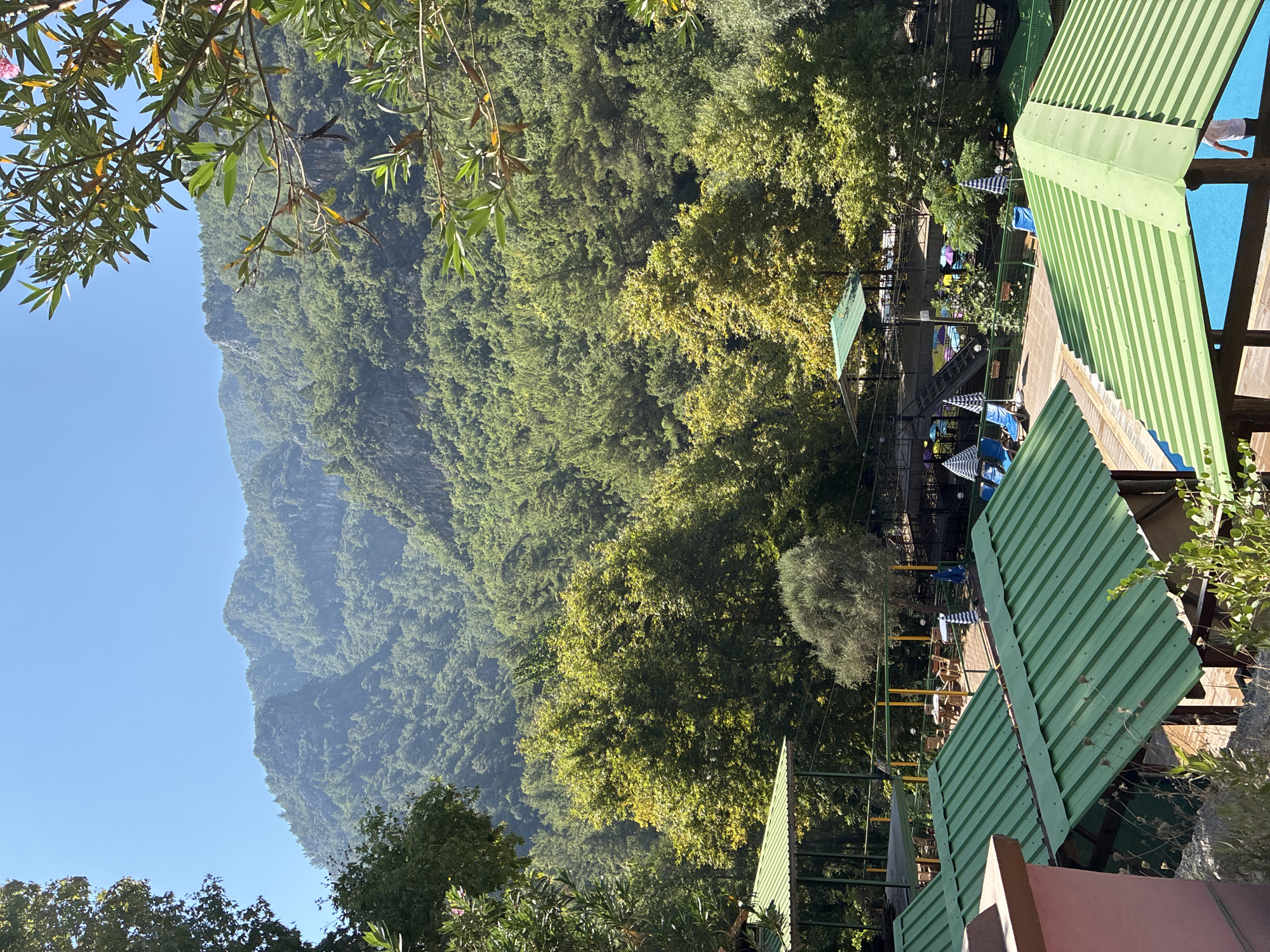 An outdoor scene is depicted with lush green mountains in the background and a resort area in the foreground. The resort features multiple green-roofed structures, lounge chairs, umbrellas, and what appears to be a swimming pool with bright blue water. Trees with dense foliage surround the area, and the sky is clear and blue. The scene suggests a warm, sunny day in a mountainous region, likely a tourist destination.