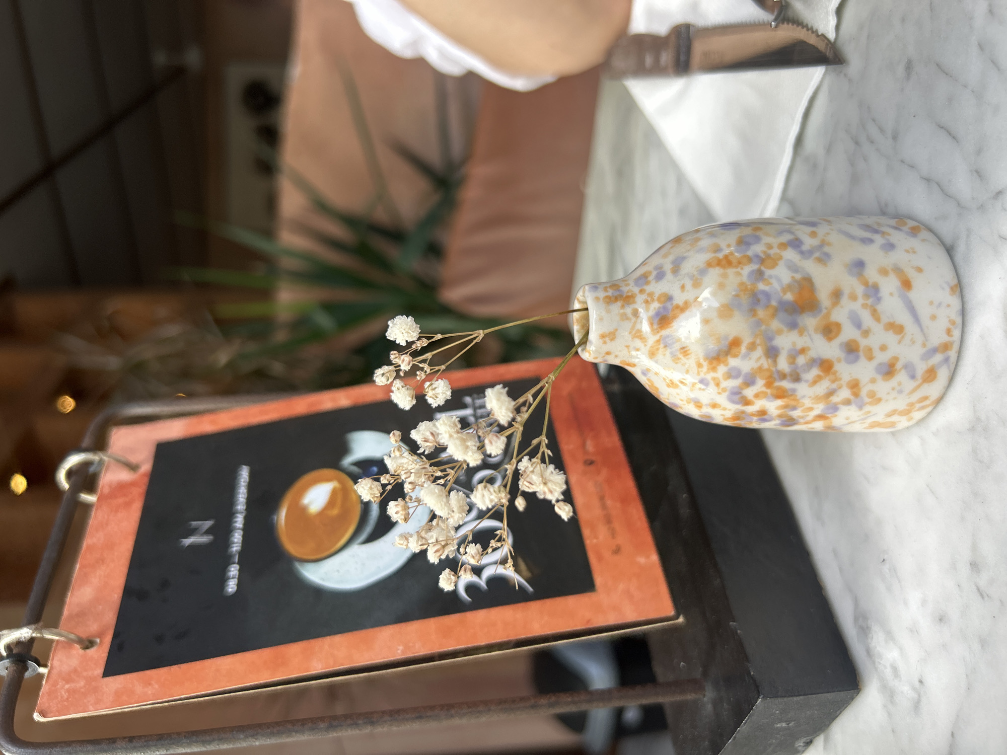A close-up shot shows a small, speckled ceramic vase with dried white flowers on a marble tabletop in Kuta, Indonesia. Behind the vase, an orange-bordered menu stands on a black stand. The menu features the letter "N" and text that reads "08:30-11:00 AM EVERYDAY" along with an image of a latte with heart art. In the background, a person's arm in a white sleeve is visible, along with out-of-focus greenery and a blurred seating area. A knife and white napkin are positioned to the right of the vase.