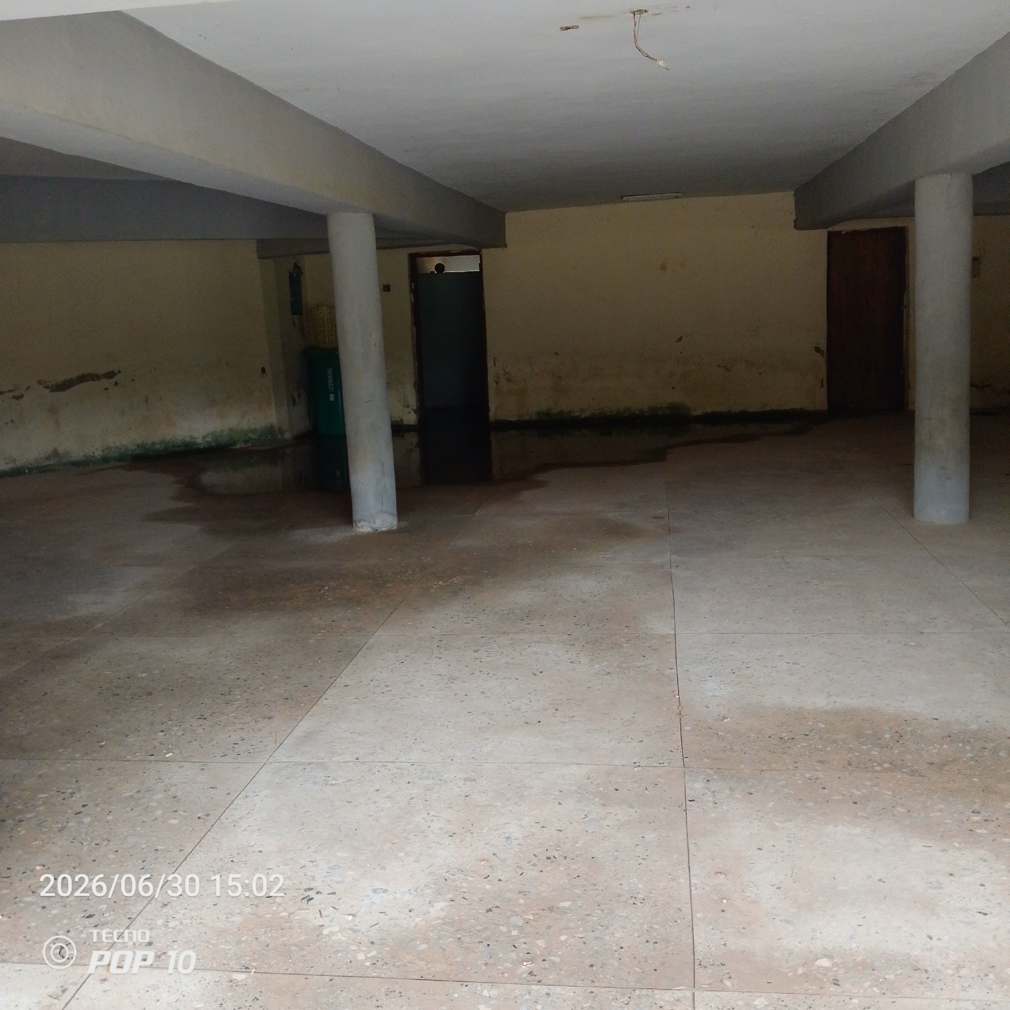 The image shows an interior view of an empty, somewhat dilapidated room, likely a part of a building under construction or in disrepair. The setting appears to be a covered outdoor space or a large, open room with concrete pillars supporting a concrete ceiling. The walls are painted a light, yellowish-tan color and show signs of moisture damage and discoloration, particularly near the floor where there are dark, damp patches and green algae growth.
The floor is made of large, square concrete tiles with a speckled pattern. There are visible water stains and puddles on the floor, suggesting recent leakage or a lack of proper drainage. In the background, there are two doorways, one on the left and one on the right. The doorway on the left is partially obscured by a green cylindrical object, possibly a trash bin or a large container, and a pillar. The doorway on the right is a dark wooden door.
The ceiling is plain concrete, and a bare wire with a light fixture dangles from it, indicating incomplete electrical work. The overall impression is one of neglect and emptiness.
There are no people visible in the image. The time of day is indicated by the timestamp "2026/06/30 15:02" in the lower left corner, which also includes the text "TECNO POP 10", suggesting it is late afternoon. There are no explicit weather cues, but the dampness indoors could imply high humidity or recent rain. The location context is Jalingo, Nigeria.