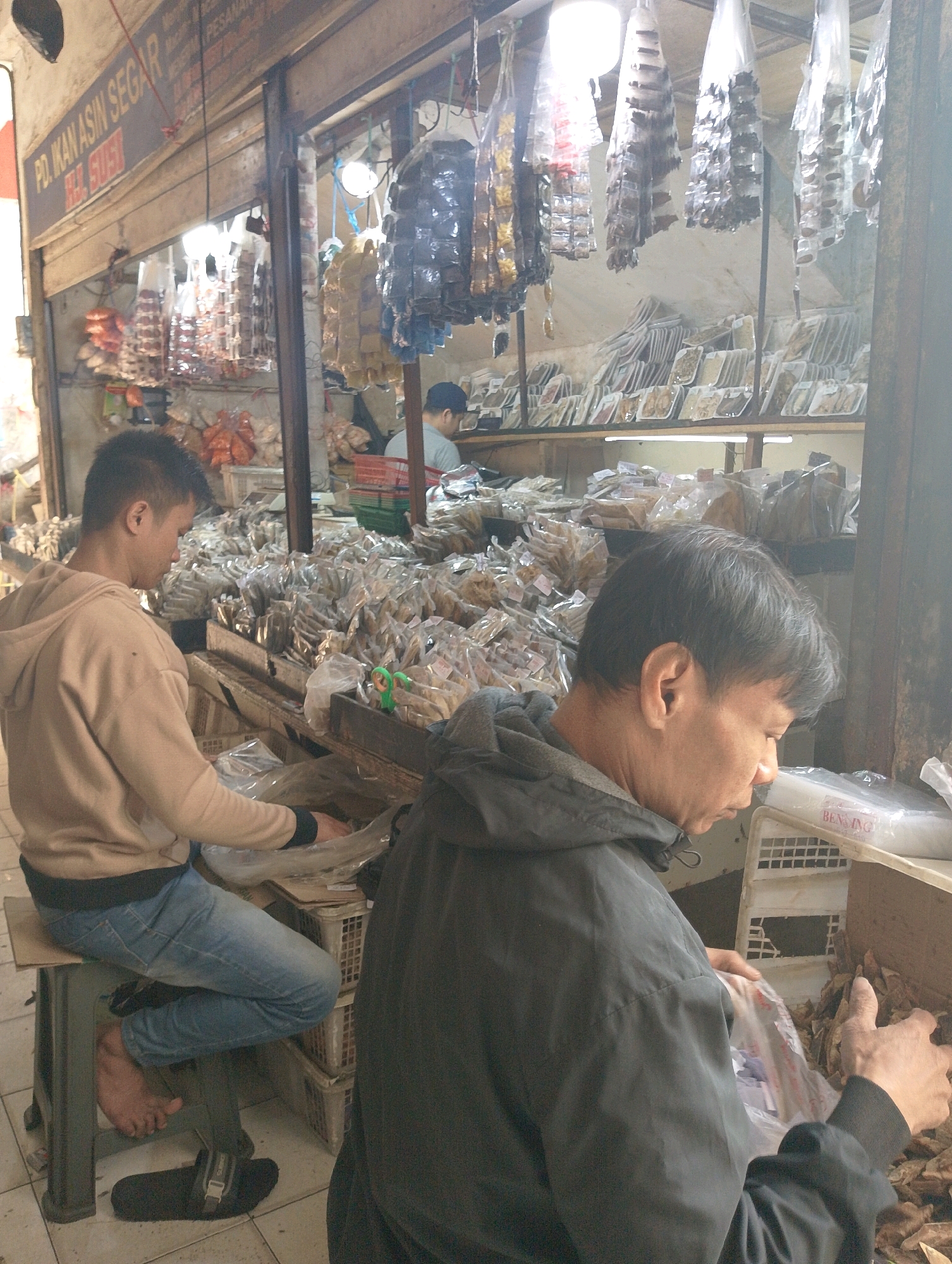 The image depicts a scene at an outdoor market stall, possibly in Lembang, Indonesia, given the signage visible. Two individuals are present. In the foreground and slightly to the right, a person wearing a dark hooded jacket is looking down and appears to be sorting through plastic-wrapped goods. Their hands are visible, and they are holding a plastic bag. To the left, another person, seen from the side and wearing a light brown hooded sweatshirt and blue jeans, is seated on a dark stool and is also looking downwards, possibly at items on the counter. Their bare foot is visible, along with a black sandal on the floor. The stall itself is laden with various products, predominantly packaged food items, many of which are transparently wrapped and appear to be dried or preserved goods. Shelves and baskets are filled with these items, and some are hanging from above. A sign with Indonesian text is visible on the left side of the stall. The environment appears to be a bustling marketplace with goods on display for sale.