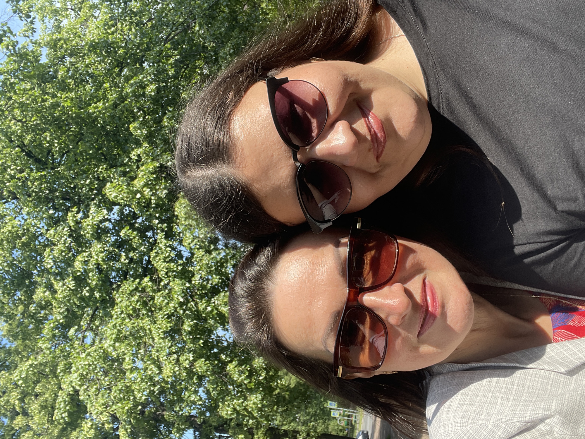Two women, likely adults, are taking a selfie outdoors on a sunny day. They are positioned close together, with their faces filling most of the frame. Both are wearing sunglasses. The woman on the left has dark hair pulled back and is wearing a light-colored, textured jacket over a colorful garment. The woman on the right has dark, long hair and is wearing a dark shirt. Their faces are turned towards the camera, and both appear to be smiling. Behind them, out of focus, is lush green foliage from trees, suggesting a park or a tree-lined street. The lighting indicates a bright, possibly warm day. The overall impression is a casual, happy moment captured outdoors.