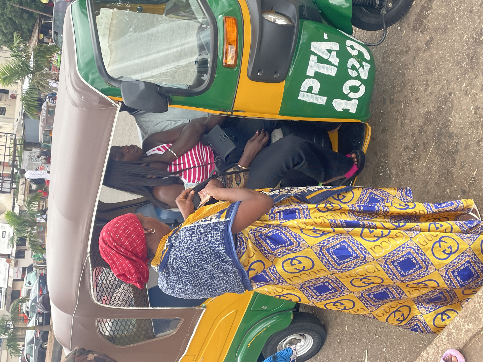 In the city of Barakin Lerawi, Nigeria, a three-wheeled auto-rickshaw, painted green and yellow, is parked on a paved surface. The rickshaw displays the license plate "PTA-1029." A woman wearing a red headscarf, a blue and white patterned scarf draped over her shoulders, and a yellow dress with blue and gold chain patterns is standing next to the rickshaw. She is holding some banknotes in her hands, possibly engaged in a transaction. Inside the rickshaw, a woman with long black hair, wearing a pink and white striped top and black pants, is seated. Another passenger, wearing a grey shirt, is also visible inside. The background shows buildings, palm trees, and other vehicles, indicating an urban setting.