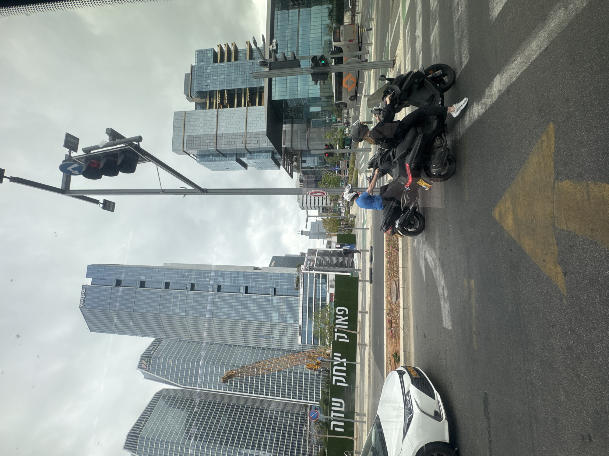 The image depicts a street scene in Tel Aviv, Israel. Modern high-rise buildings dominate the background. A construction crane is visible near one of the buildings. In the foreground, two people on electric scooters are stopped at a traffic light. The traffic lights show red and green signals. A white car is partially visible in the lower right corner of the frame. A green sign along the road barrier contains writing in Hebrew. The sky is overcast.
