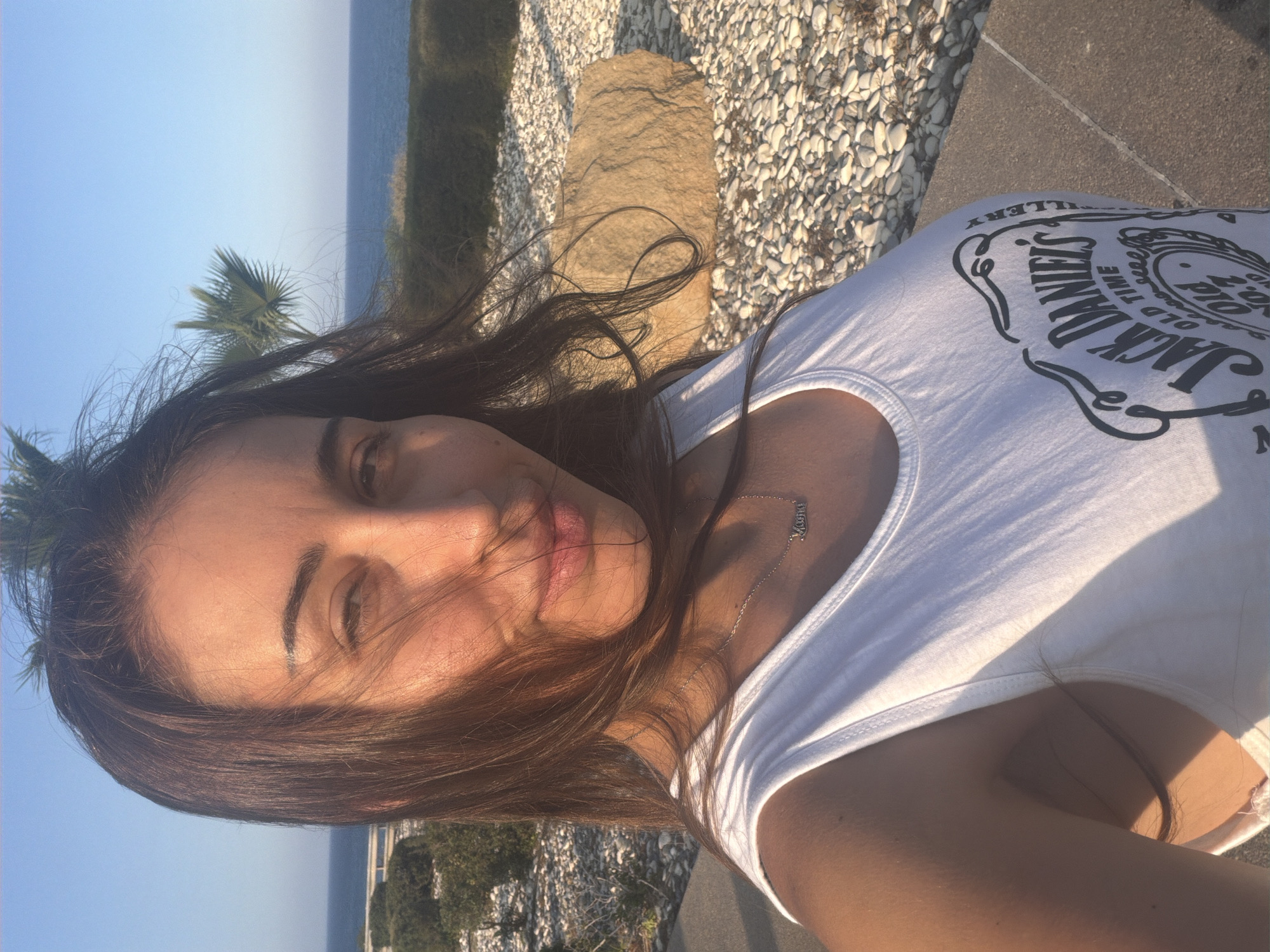A woman with long, dark hair is taking a selfie outdoors. She is wearing a white tank top with a "Jack Daniel's Old Time Old No. 7 Distillery" logo. She has a delicate silver necklace with a pendant that appears to say "Mama". The background shows the sea, a palm tree, and a rocky area. The lighting suggests it is either early morning or late afternoon, with the sun casting warm tones on her face and skin. The location is identified as Meneou, Cyprus.