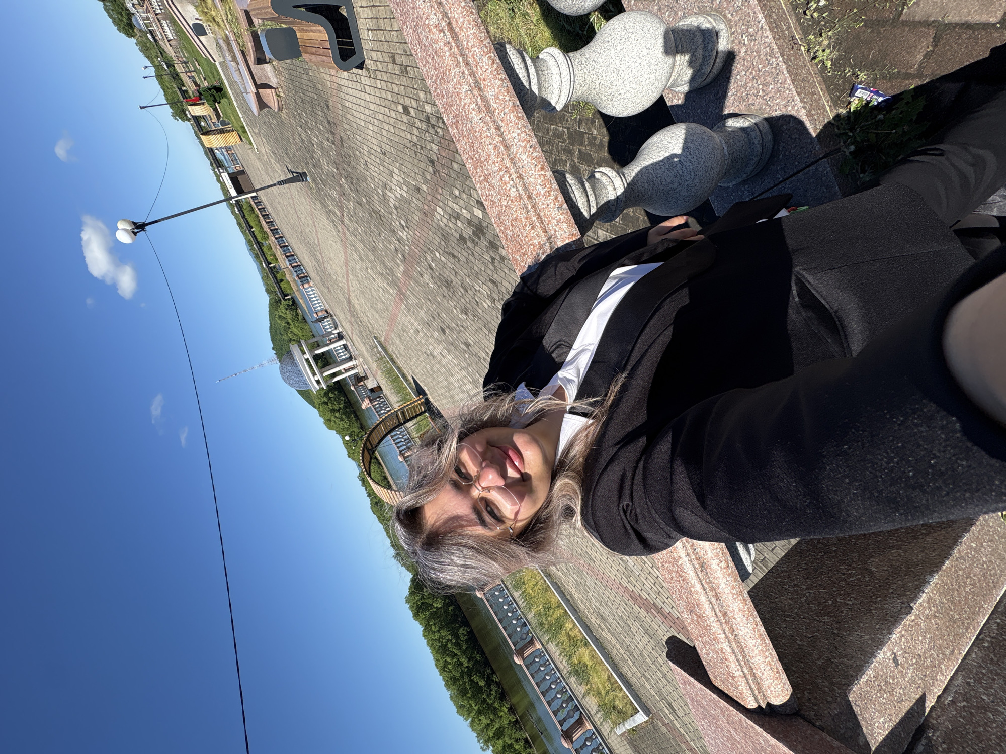 A woman takes a close-up selfie in a park in Birobidzhan, Russia, on a bright, sunny day. She is positioned to the right of the frame, looking directly at the camera with a playful smile and her tongue slightly sticking out. She has long, wavy, light-colored hair, wears glasses, a black blazer, and a white shirt.
Behind her, the scene extends to the left. A paved promenade with light-colored bricks stretches into the distance, lined with benches and lampposts. To the right of the woman, a decorative low wall or railing, made of a speckled reddish-brown stone, features large, gray, chess piece-like ornaments. Strong shadows from these objects and the woman indicate direct sunlight.
Beyond the promenade, a body of water (likely a river or canal) is visible, with green trees lining its distant bank. A distinctive circular or domed structure is partially visible further down the waterway. The sky is clear and blue with a few scattered white clouds. A thin black wire or cable stretches horizontally across the upper portion of the sky. A small piece of blue and white trash is visible on the ground near the base of the decorative wall. The overall impression is one of a cheerful and relaxed outdoor moment in a public park setting. No legible text is visible.