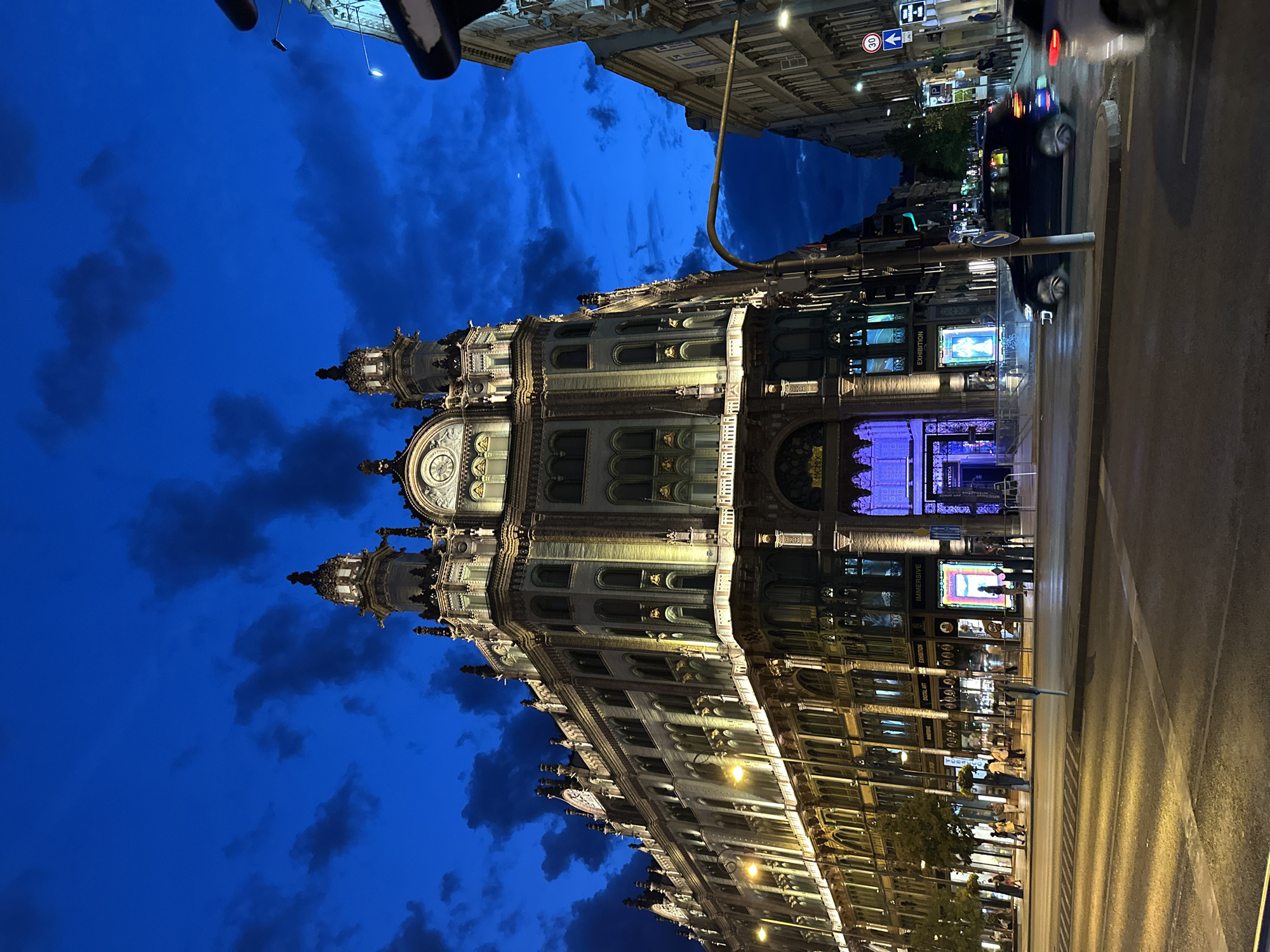 The image depicts the Thonet House in Budapest, Hungary, at dusk or early evening. The ornate building, illuminated by warm lights, features classical architectural elements, including arched windows, decorative carvings, and turrets. The sky is a deep blue, with dramatic dark clouds suggesting recent or impending rain. In the foreground, a street intersection with traffic lights and road markings is visible. A dark-colored car is in motion, blurred due to the long exposure. Pedestrians are walking along the sidewalk in front of the building, which houses shops and what appears to be an exhibition space with brightly lit displays. The overall scene captures the grandeur of Budapest's architecture at night, with the contrast between the illuminated building and the dark sky.