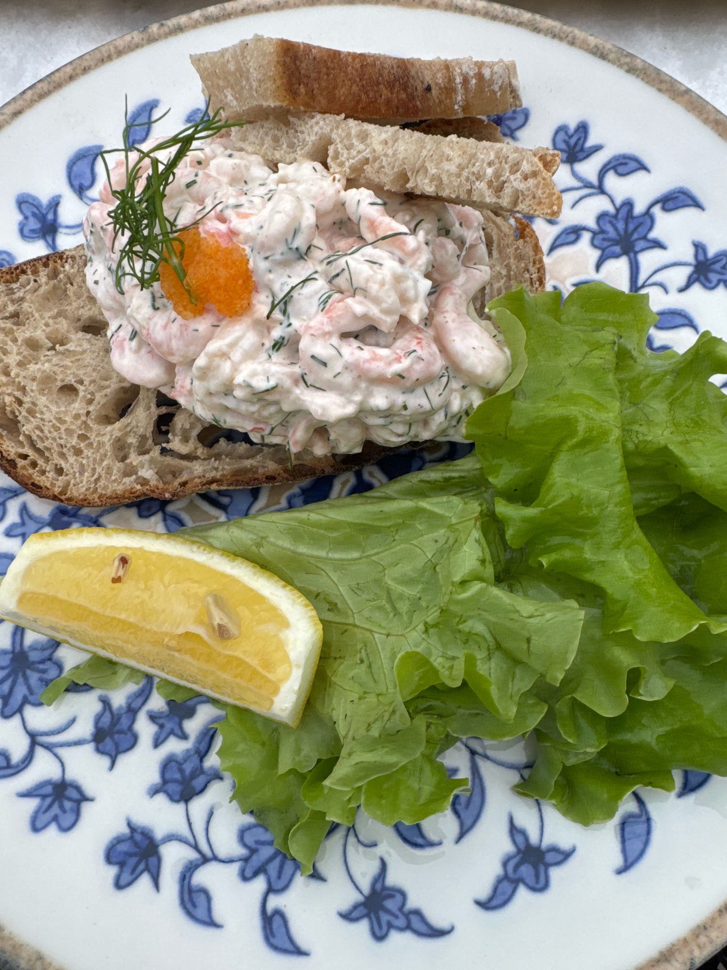 A close-up, overhead shot shows a shrimp salad open-faced sandwich on a blue and white floral plate. The sandwich consists of two slices of dark rye bread, one partially obscured by the other. The top slice is covered with a generous portion of shrimp salad, a creamy mixture with finely chopped dill and small orange roe. A sprig of fresh dill garnishes the salad, along with a small mound of bright orange roe. To the right of the sandwich are several crisp green lettuce leaves. A wedge of bright yellow lemon rests on the lettuce, its skin showing some texture and a seed visible within. The plate's rim has a speckled brown edge. The overall scene suggests a light meal served outdoors, likely on a sunny day, given the bright lighting and natural colors. There are no people or animals present. The setting appears to be a cafe or restaurant in Vaxholm, Sweden, a city known for its picturesque archipelago setting and waterfront dining options.