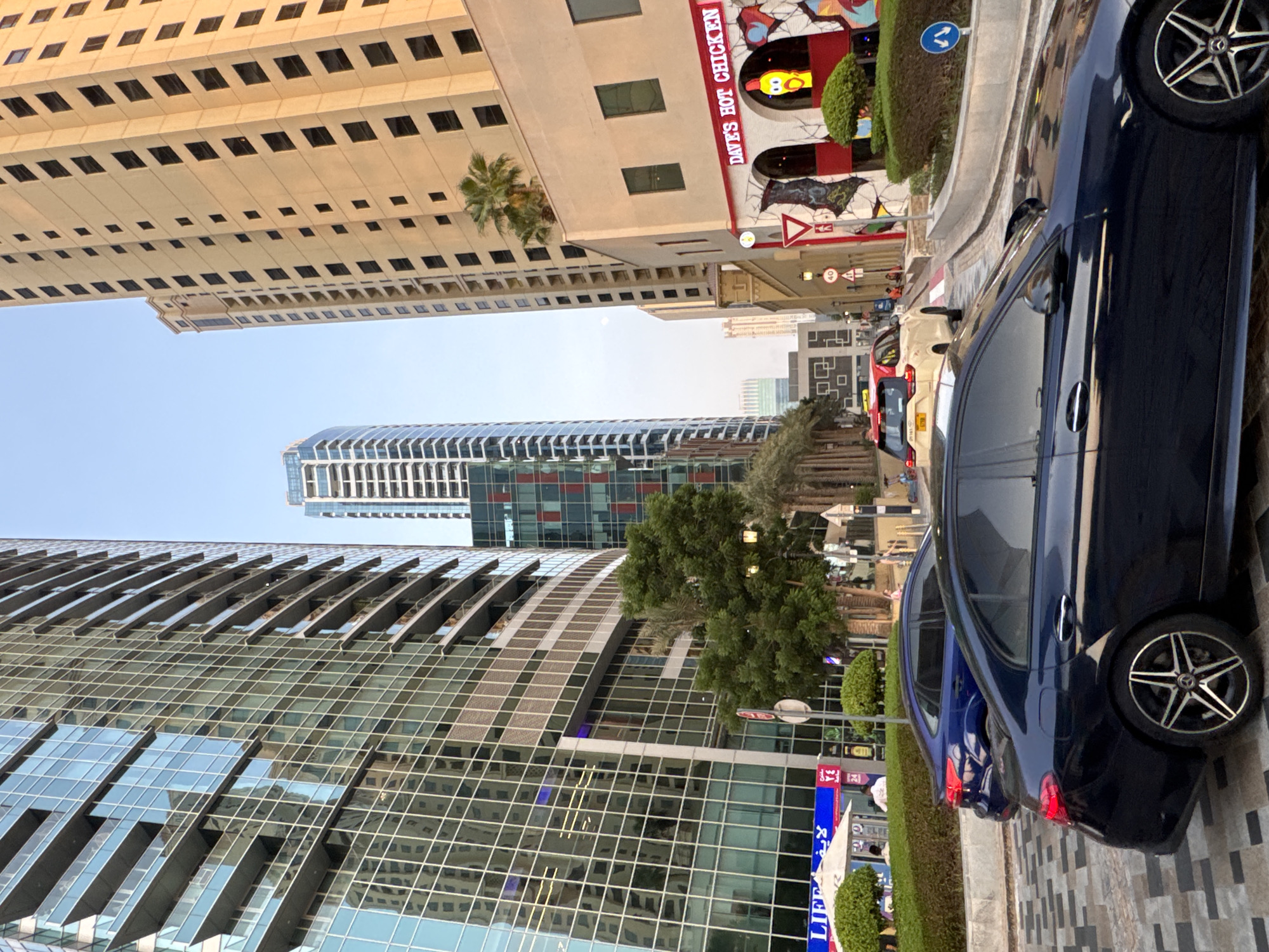 The image depicts a street scene in Dubai, United Arab Emirates, likely in a modern urban district such as Dubai Marina or Jumeirah Beach Residence, characterized by tall, contemporary skyscrapers. In the foreground, a dark blue Mercedes-Benz sedan is parked. Behind it, a blue sedan is also visible. A yellow and white taxi is driving away from the camera on the right side of the street. The buildings feature glass facades and balconies, reflecting the clear sky and surrounding structures. On the right side of the image, a building with a red and white facade displays signage for "DAVE'S HOT CHICKEN" and a cartoonish depiction of a chicken. The street is paved with interlocking tiles, and there is a landscaped area with bushes and palm trees bordering the sidewalk. Several traffic signs are present, including a speed limit sign for 40 and directional signs. The sky is clear with a soft, warm light, suggesting it is either early morning or late afternoon. The overall environment is clean and well-maintained, consistent with the urban landscape of Dubai.