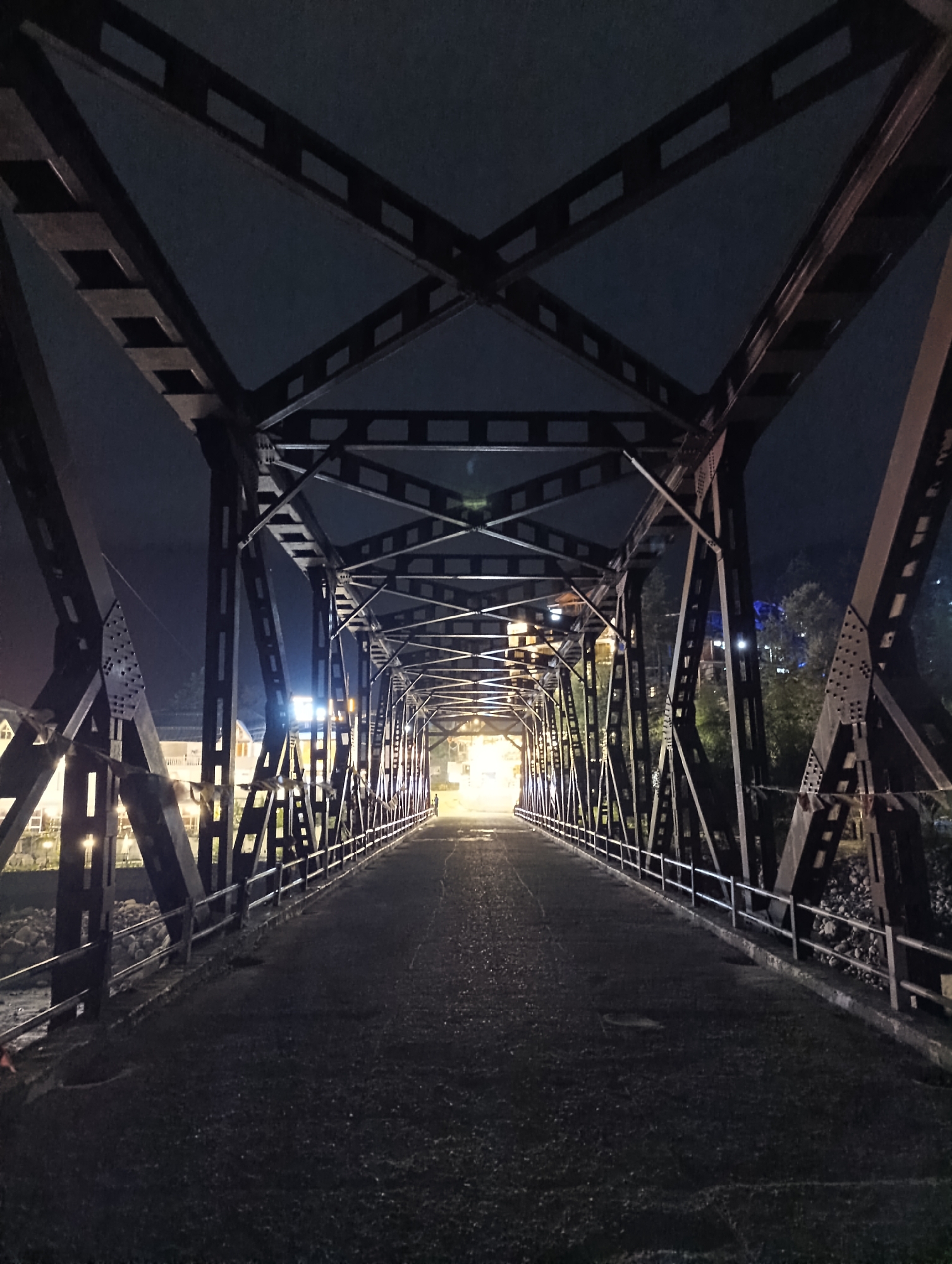 The image depicts a dimly lit, empty steel truss bridge at night, situated in the city of Kulang, India. The bridge's structure, characterized by its intricate network of beams and supports, dominates the foreground and midground, creating a tunnel-like effect. The roadway surface appears worn and dark, with reflections of distant lights visible. The sky above is dark and clear. On either side of the bridge, structures and trees are faintly illuminated by artificial lights, suggesting an urban or semi-urban environment. No people are visible on the bridge.