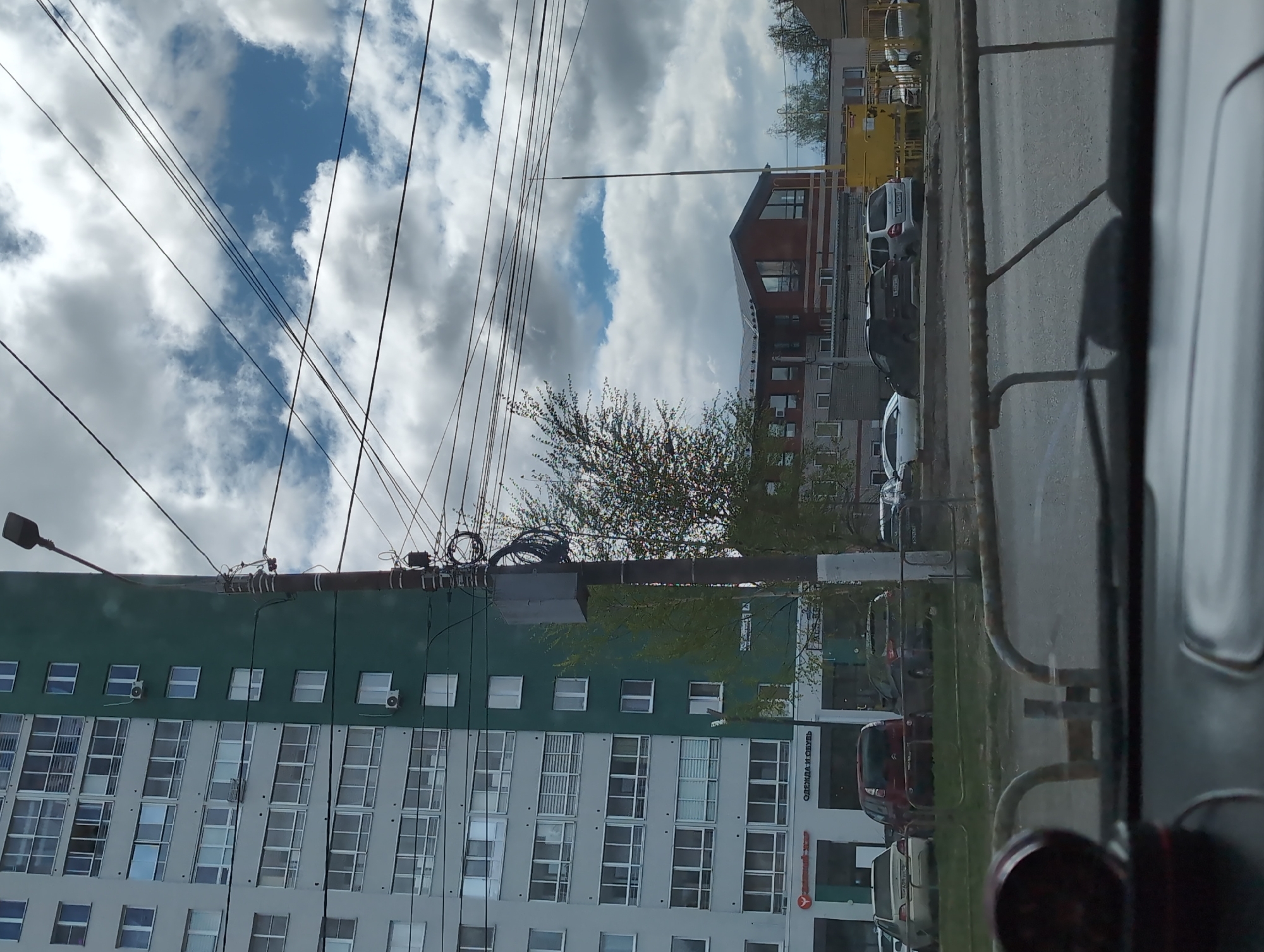 The image shows a view from inside a vehicle looking out onto a street in Izhevsk, Russia. A tall apartment building with a green and white facade dominates the left side of the frame. Power lines are visible against the sky, which features a mix of blue sky and white clouds. A building with a distinctive brown roof is also in view. On the right side of the frame, a road and parked cars are partially obscured by a metal railing. A tree with new leaves is located in the mid-ground. The words "ОДЕЖДА И ОБУВЬ" are visible on a building.
