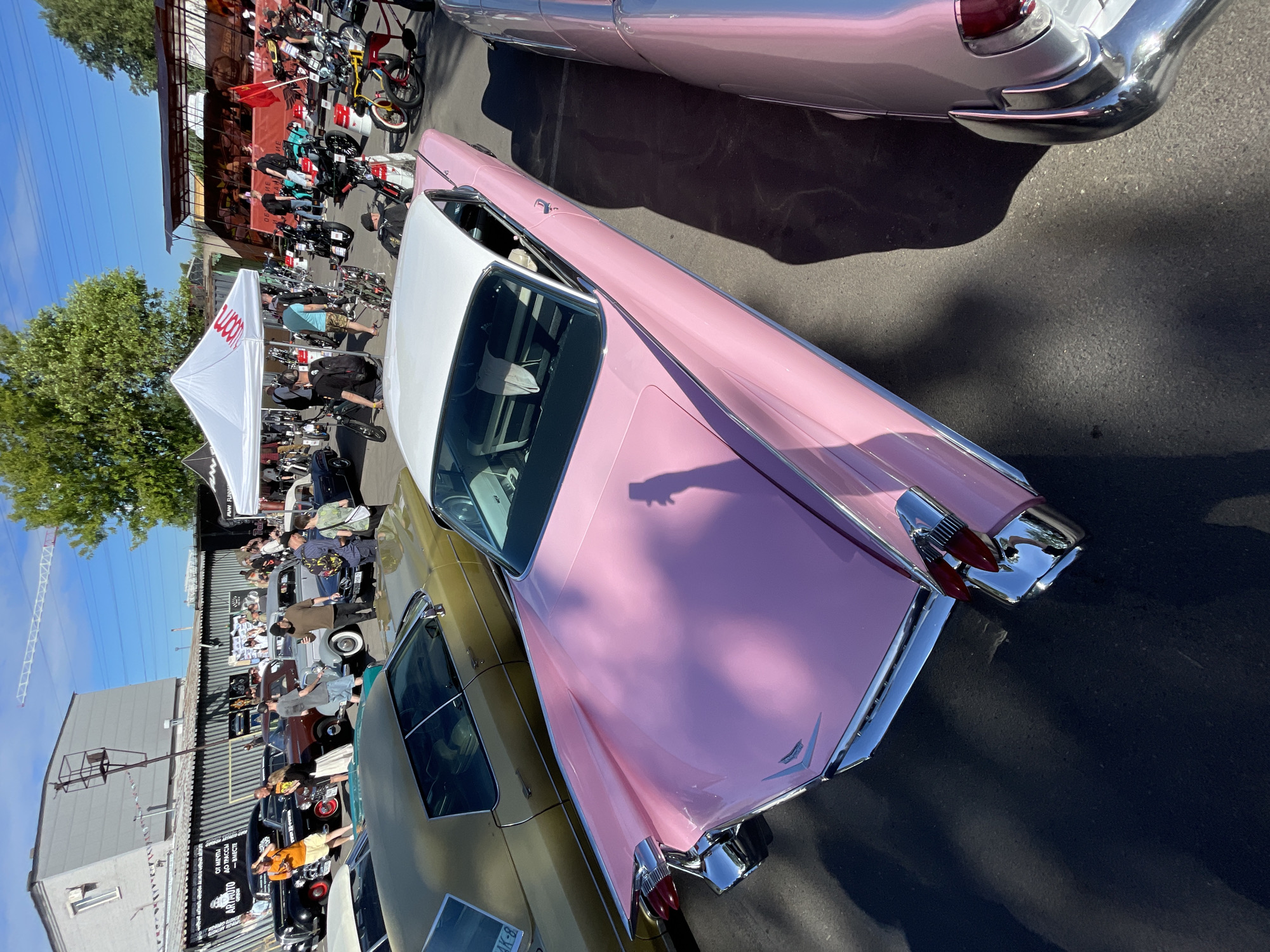 The image depicts an outdoor car show event in Saint Petersburg, Russia, under a clear blue sky. In the foreground, a vintage pink Cadillac with a white roof is prominently displayed. Its distinctive tail fins and chrome accents are visible. To the left of the pink Cadillac, a gold-colored vintage car is parked. The background reveals a gathering of people, various other classic cars, and a white tent with branding. Further back, a stage area with seating is visible, along with more attendees. The setting appears to be an urban outdoor space, possibly a parking lot or an open area adjacent to industrial buildings, suggested by the presence of a large gray building with a window and a gantry on its roof. The overall atmosphere suggests a social gathering focused on vintage automobiles.
