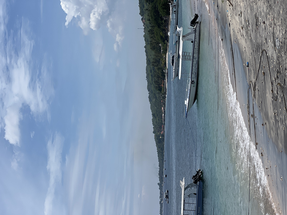 The image depicts a coastal scene with several boats anchored close to a sandy beach. Clear turquoise water separates the boats from the shore. Lush green vegetation covers the land extending along the coastline. The sky is a mix of light blue with scattered white clouds. The image is taken during daytime, under bright sunlight.