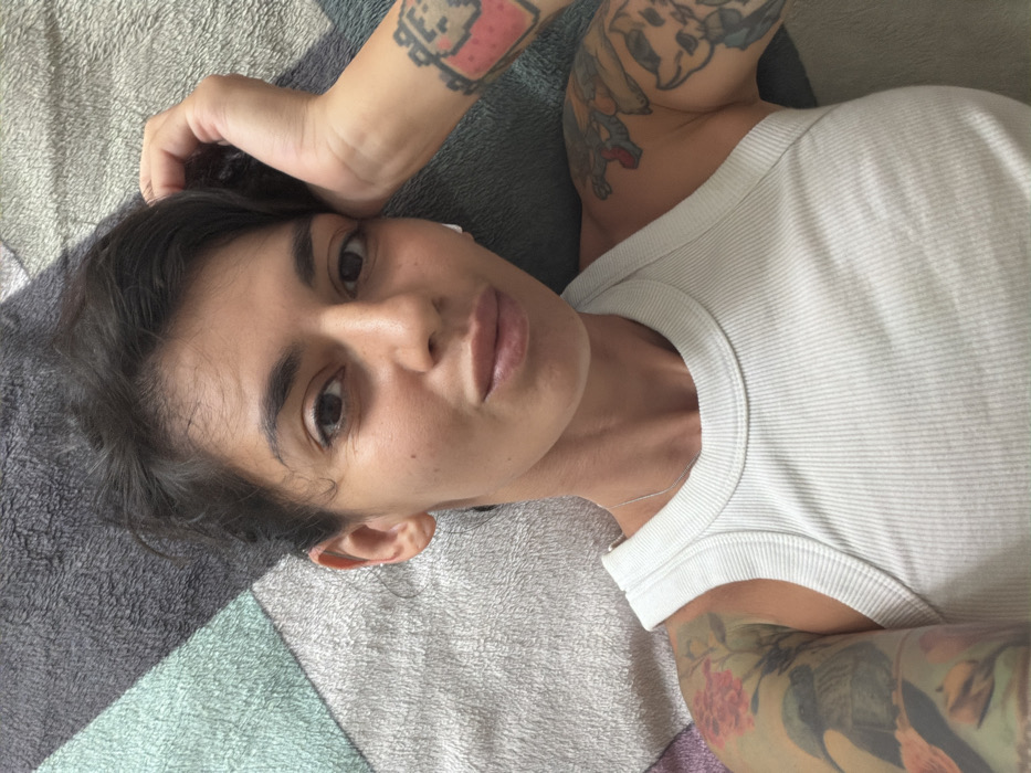 A female individual is depicted in a close-up, overhead shot, likely lying on a bed or soft surface, in Nusa Dua, Indonesia. She possesses a medium skin tone, dark, short, disheveled hair, and is looking directly at the camera with a neutral expression, her lips slightly parted. A white earbud is visible in her right ear. She wears a light-colored, ribbed tank top and a thin silver chain necklace. Both arms feature extensive tattoos: the left arm displays a colorful sleeve with a bird on a branch amidst pink flowers and green foliage, while the right arm exhibits a sleeve with a pixelated character on the forearm and a stylized animal skull or head along with other designs further up the arm. Her right hand is positioned near or on her head, with fingers partially obscured by her hair. The background is a soft, textured fabric, potentially bedding, displaying a geometric pattern in shades of grey, light blue, and off-white.