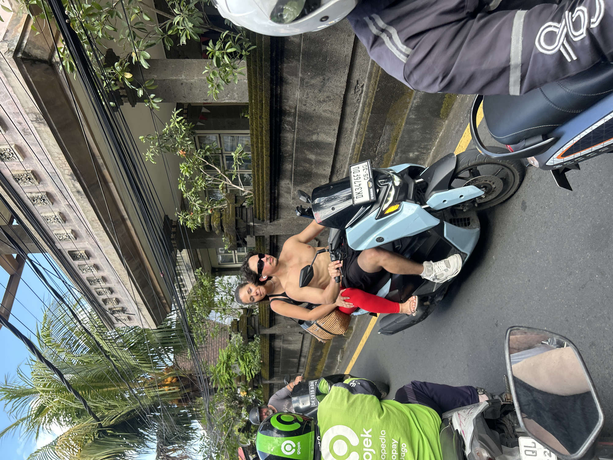 A man and woman ride on a scooter in traffic in North Kuta, Indonesia. The man is shirtless and wearing black shorts and white sneakers. The woman is wearing a black tank top and red leggings. They are behind a person on a green scooter with a Gojek logo. A building with intricate stone carvings is visible in the background, along with palm trees and power lines. The sky is blue and clear. The scooter has a license plate that reads "DK3476FDU 05-36".