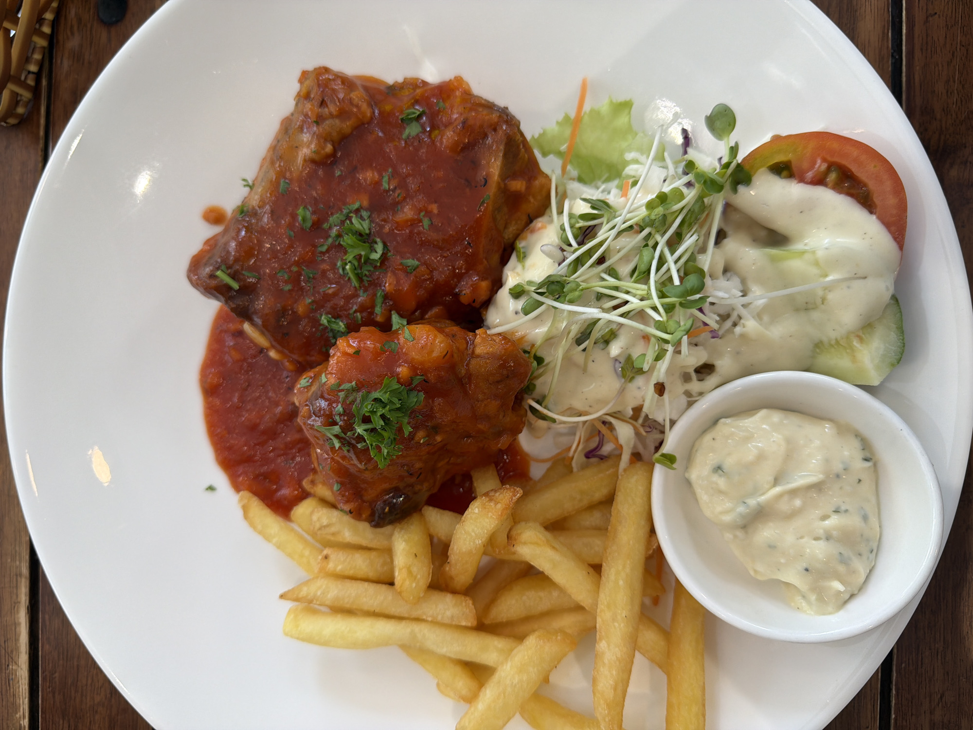 A white plate holds a meal of ribs, french fries, salad, and a small dish of sauce. The ribs are covered in a red sauce and sprinkled with green herbs. The french fries are golden brown and piled next to the ribs. The salad is a mixture of lettuce, shredded carrots, and sprouts, topped with a creamy dressing. The sauce dish contains a light-colored, creamy sauce with herbs. The plate is set on a wooden table. The meal appears to be in a restaurant in Nha Trang Ward, Vietnam.