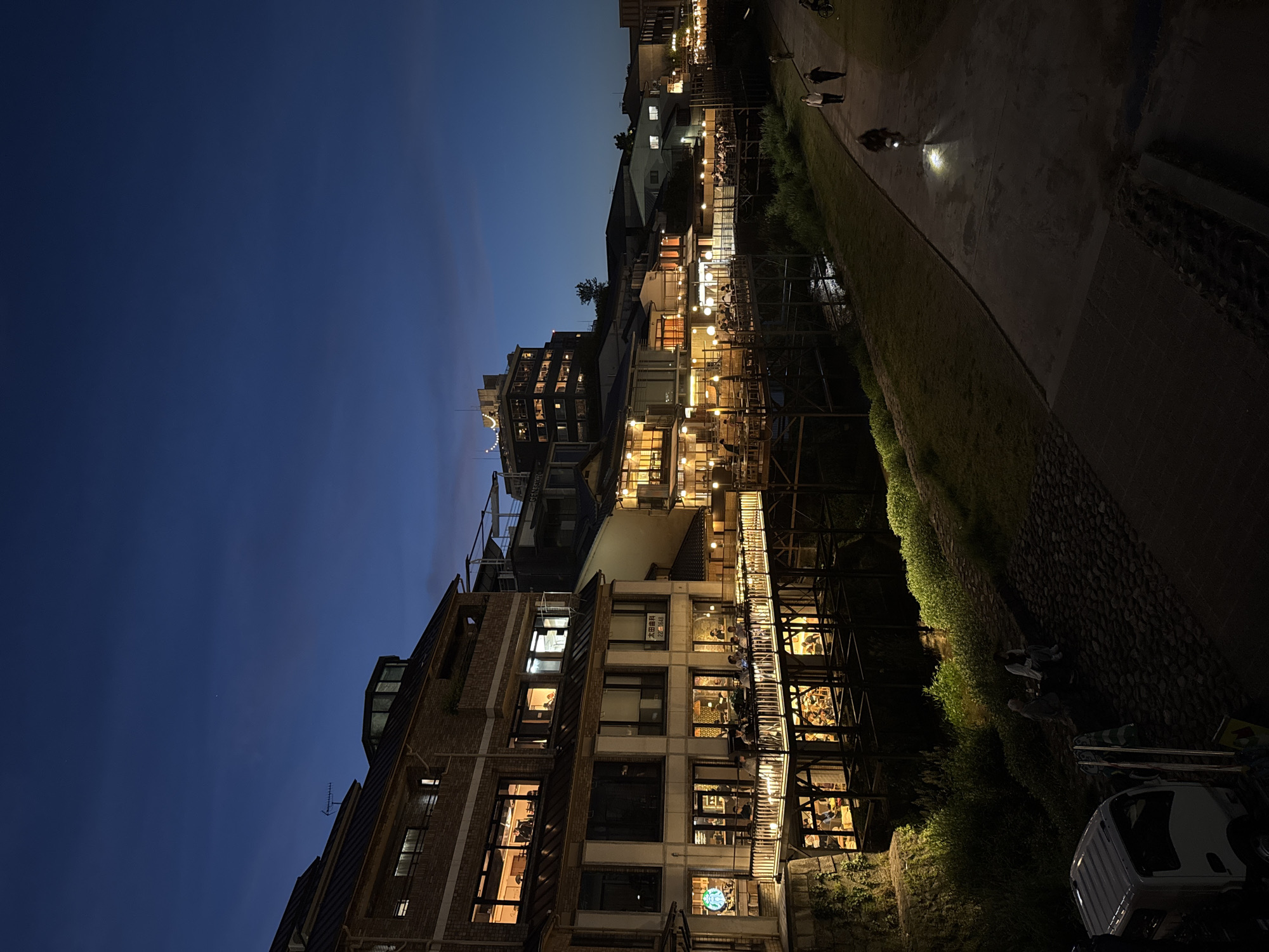 The image shows a nighttime scene in Kyoto, Japan, with a river or canal running alongside buildings and a path. The buildings are multi-storied with illuminated windows, suggesting commercial or residential use. Some structures appear to be on multiple levels, possibly with outdoor seating areas along the water. People are visible on the path along the river, including a cyclist and walkers. The overall environment is urban, illuminated by streetlights and building lights. The time of day is late evening or night.