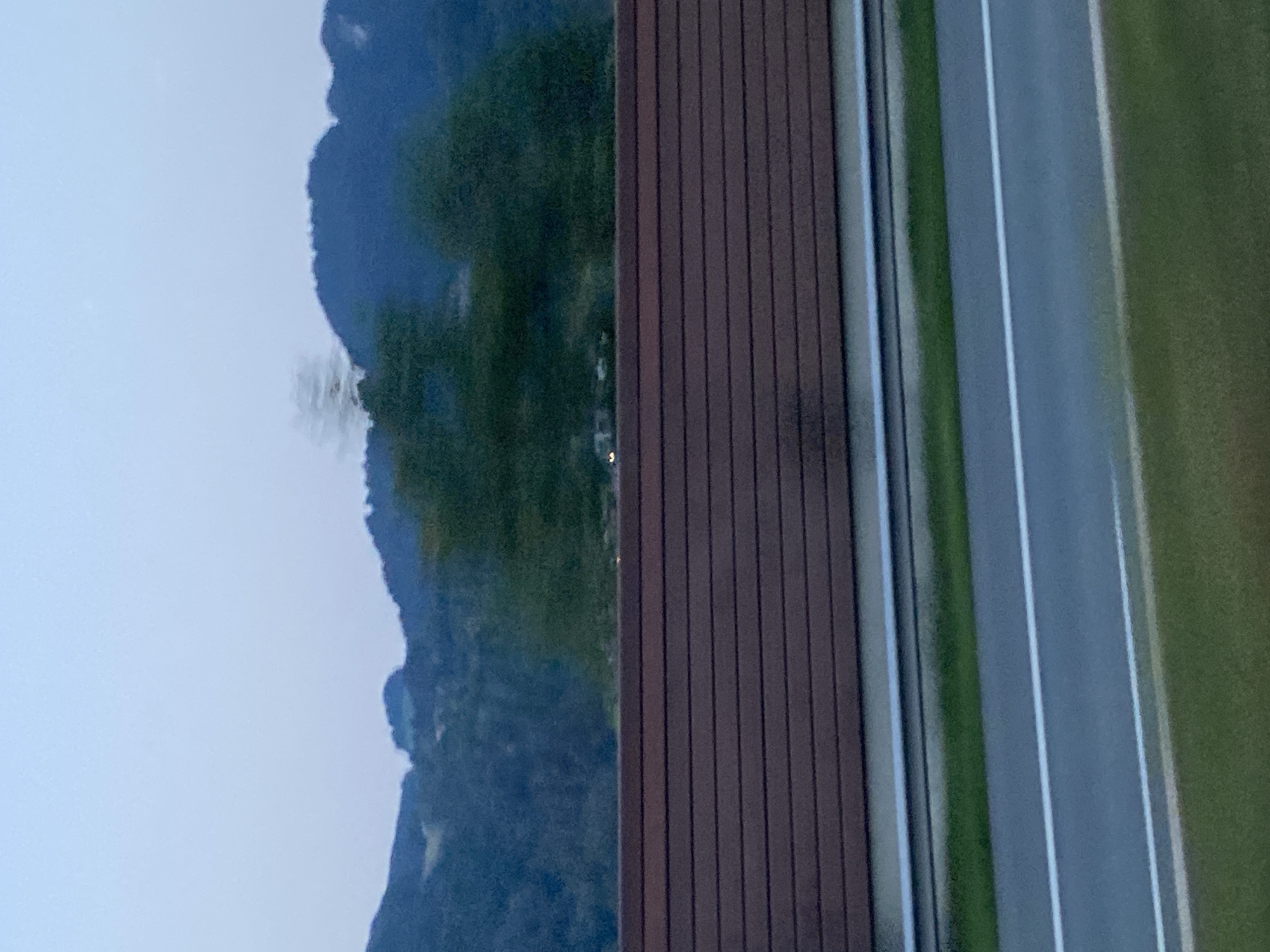 The image depicts a view from a vehicle, likely on a highway, looking towards a treeline and distant mountains under a hazy, pale blue sky. A dark brown, horizontally-slatted noise barrier runs across the middle ground, obscuring the view behind it. Below the barrier, a paved road with white lane markings is visible, bordered by a strip of green grass. The overall impression is one of travel or transit, with the background elements slightly blurred due to motion. The location is identified as Oberaudorf, Germany, suggesting a highway passing through or near this Bavarian town. There are no people or discernible animals in the image. The scene appears to be captured during twilight or overcast conditions, given the muted lighting.