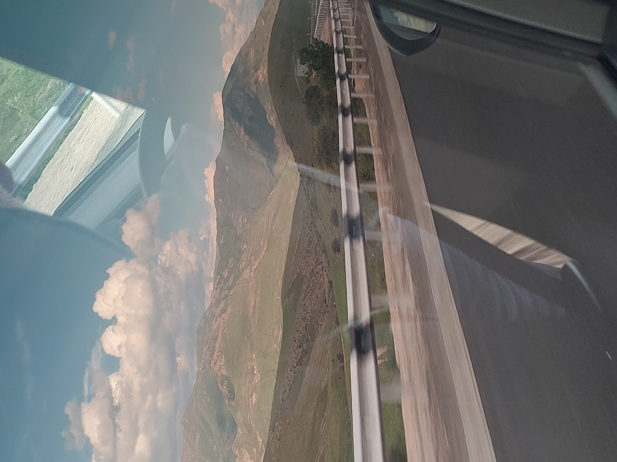 The image captures a vast mountainous landscape viewed from inside a moving vehicle, likely a car. The primary scene features a paved road on the right side, extending into the distance, bordered by a multi-bar metal safety barrier. To the left of the road, a series of large, undulating mountains dominate the mid-ground and background. These mountains appear to be covered in sparse green and brown vegetation, suggesting a dry environment or a specific season. A small building or structure is faintly visible nestled among trees on one of the distant hills.
The sky, occupying the upper left portion of the image, is a clear blue, adorned with large, fluffy white cumulus clouds. The time of day is daytime, with ample natural light illuminating the scene, casting soft shadows on the mountains and clouds.
In the immediate foreground, elements of the car from which the photo was taken are visible, including a portion of a side mirror and the window frame in the upper right. A prominent reflection on the car's window in the lower left shows what appears to be another section of road and green grass, indicating the perspective from within the vehicle. The slight blur on the road barrier hints at the vehicle's movement.
The setting evokes a journey through a rural, mountainous region. Based on the provided context, the scene is located in or near Yelpin, Armenia. No people or visible text are present in the image.