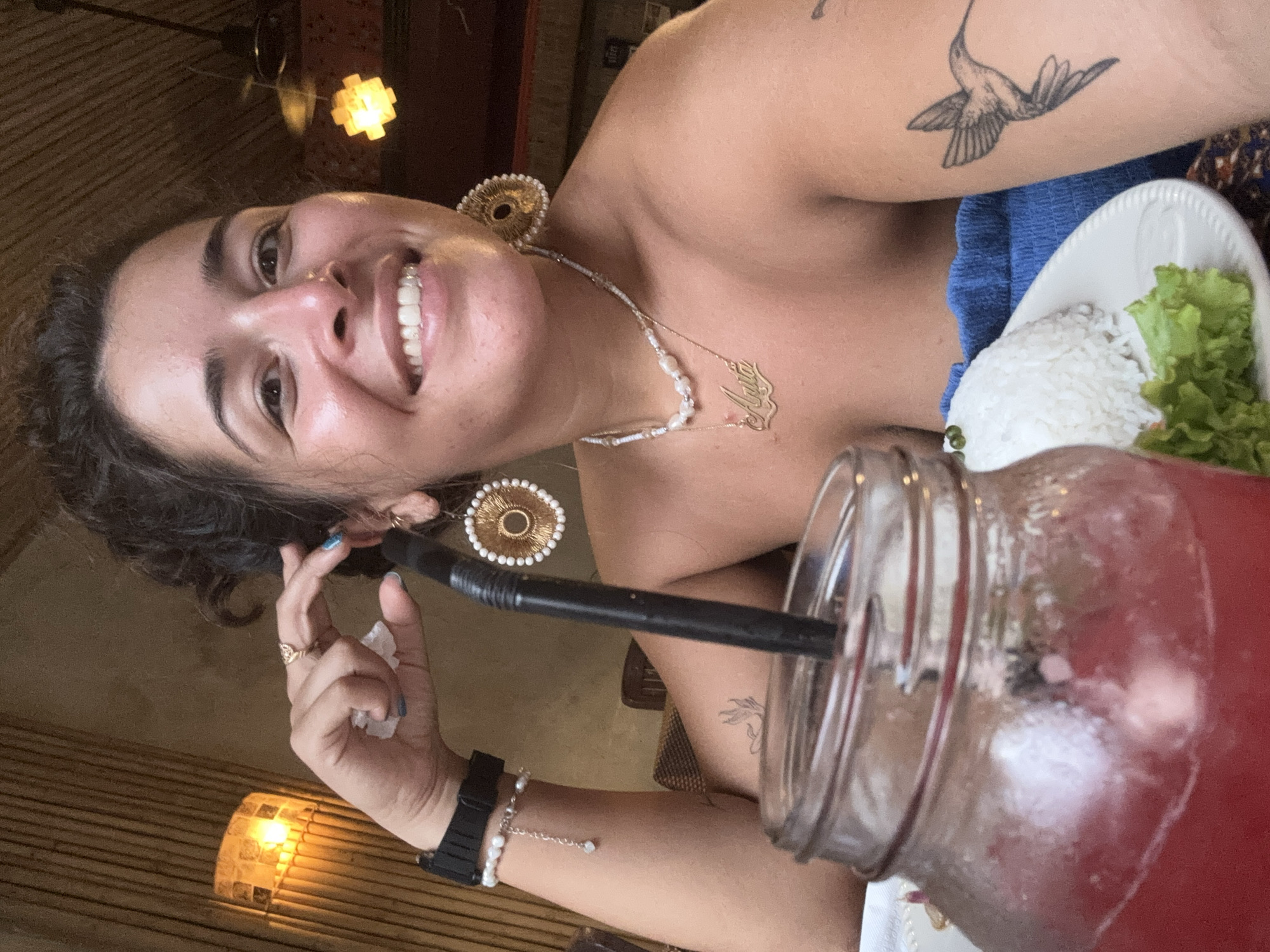 A woman with dark, curly hair pulled back sits at a table with a drink in a mason jar. She wears large, circular earrings with gold accents and white beads, a pearl necklace, and a gold nameplate necklace. A black watch is on her left wrist, and a bracelet with a small charm hangs from it. She has a hummingbird tattoo on her right arm. In front of her is a white plate with a mound of white rice and a leaf of lettuce. The background appears to be a restaurant with bamboo walls and warm lighting. The woman is smiling and holding a piece of tissue near her face. The scene is likely in Kampot, Cambodia.