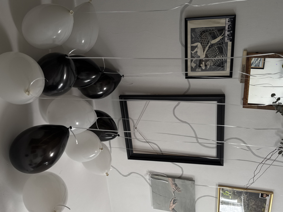 An interior scene in Tel Aviv-Yafo, Israel, displays a white-walled room decorated with multiple black and white helium balloons floating near the ceiling. Long, thin silver ribbons extend downwards from the balloons, interacting with the wall decorations. A large, empty black rectangular picture frame hangs vertically in the center of the wall. To its left, a rectangular canvas print features a grayscale rendition of the hands from Michelangelo's "The Creation of Adam." Below this, a smaller golden-framed picture shows a landscape image with two small figures in its lower right corner. To the right of the central empty frame, a framed black and white Art Nouveau style print depicts a reclining female figure on an patterned surface, with a horizontal strip of smaller framed images beneath it. Further down on the right, a wooden-framed mirror partially reflects an interior space and some greenery. Wavy gray lines are painted or applied directly onto the white walls, winding around the various framed artworks. Dry foliage or branches are visible at the bottom center, likely part of a floral arrangement. No individuals are present in the scene.