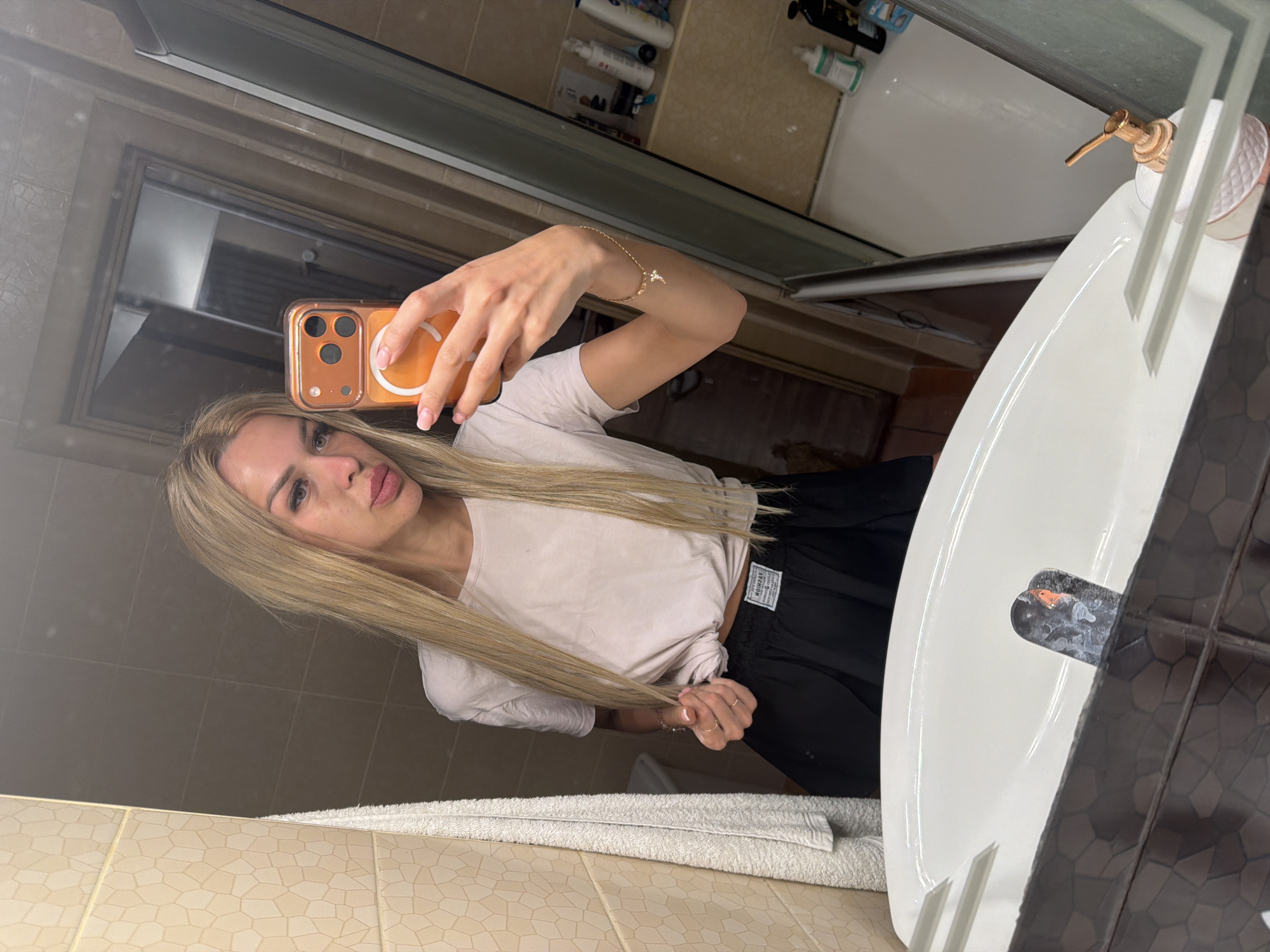 A woman with long blonde hair takes a selfie in a bathroom mirror. She is wearing a white t-shirt and black shorts. Her right hand is holding a phone with an orange case, and her left hand is pulling on her hair. She has on light makeup and jewelry on her wrist. The bathroom appears to be modern with tiled walls and a white sink. A soap dispenser with a gold pump is visible on the sink. The lighting suggests it is daytime. There is a white towel folded on the counter in front of the sink. The overall impression is a casual, everyday moment captured in a personal space.