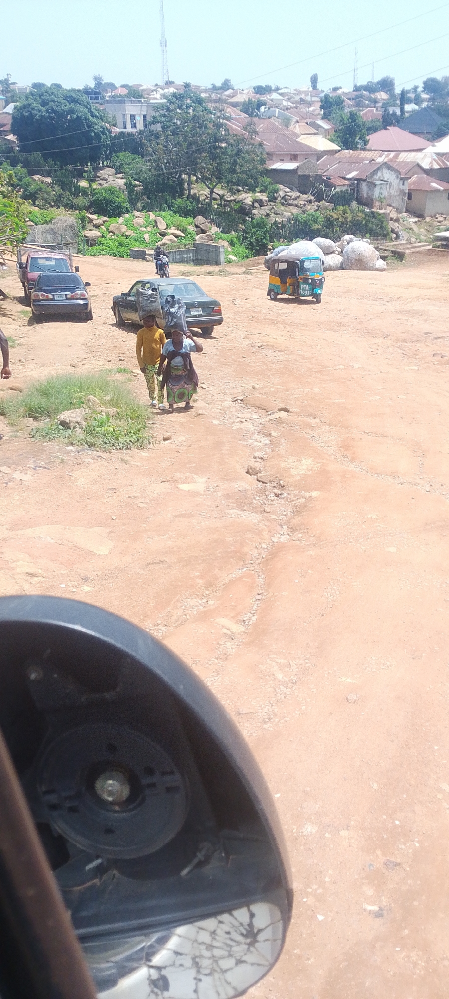 The image depicts a dusty, unpaved road in the city of Jos, Nigeria. In the foreground, the cracked side mirror of a vehicle is visible. The road is lined with a variety of vehicles, including a red truck, a dark sedan, and a three-wheeled auto-rickshaw (keke napep). Two individuals, appearing to be a woman and a child, are walking on the road, with the woman carrying a bundle on her head. In the background, a densely populated residential area with numerous houses and buildings is visible, some of which are situated on rocky terrain. A cell tower can be seen in the far distance. The overall scene suggests a typical day in a busy, developing urban environment.