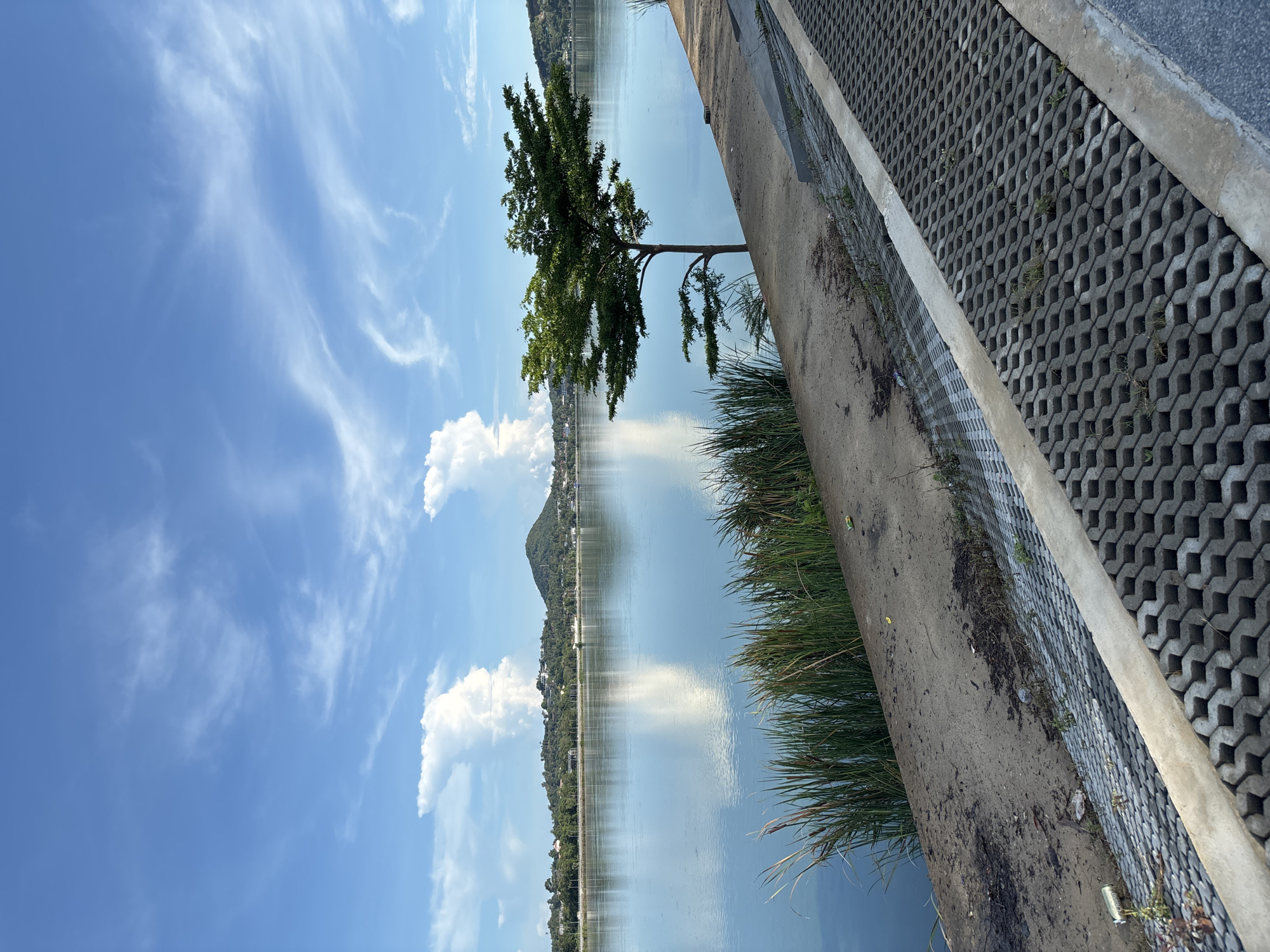 The image shows a lake in Baan Chaweng, Thailand, likely taken from a waterfront walkway or embankment. The sky is blue with scattered white clouds reflected in the calm water. A small mountain or hill is visible in the distance across the lake. A tree grows near the water's edge. The edge of the walkway is constructed with interlocking concrete blocks, and tall grass grows between the walkway and the lake. Debris and litter are visible near the walkway.