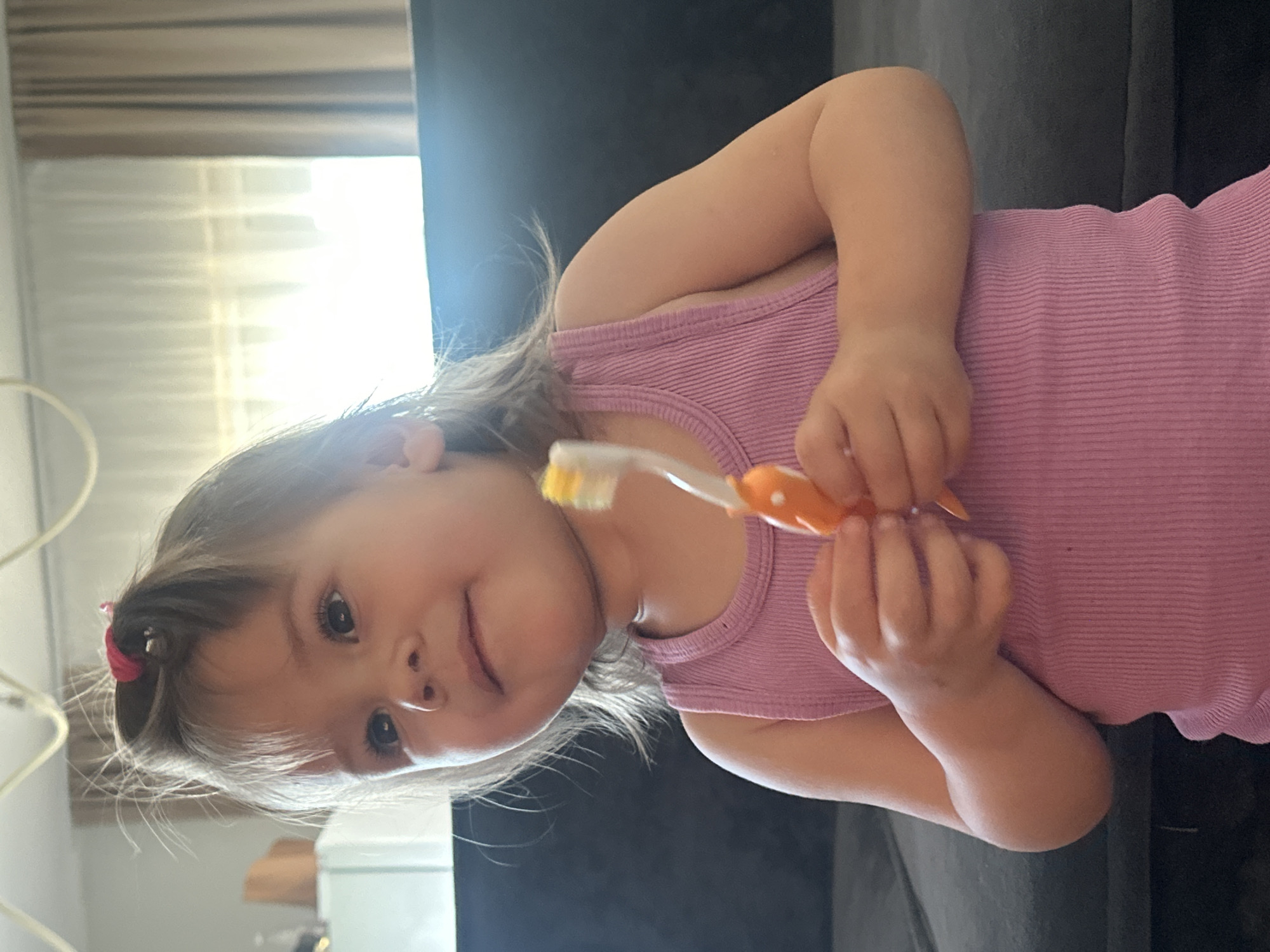 A young child, likely a girl, is featured in a close-up, chest-up shot taken indoors in what appears to be a home setting, possibly in Kharnavy, Russia. The child has light brown hair, partially tied back with a small red hair accessory, and is wearing a pink ribbed tank top. She is looking directly at the camera with a gentle, soft smile.
The child is holding a toothbrush with an orange handle and yellow and white bristles. Her small hands are wrapped around the toothbrush. In the background, bright natural light streams in from a window, which has visible horizontal blinds, indicating it is daytime. A dark sofa or similar furniture item is visible behind the child. The focus is primarily on the child, with the background slightly blurred. There is no visible text in the image. The child appears calm and content.