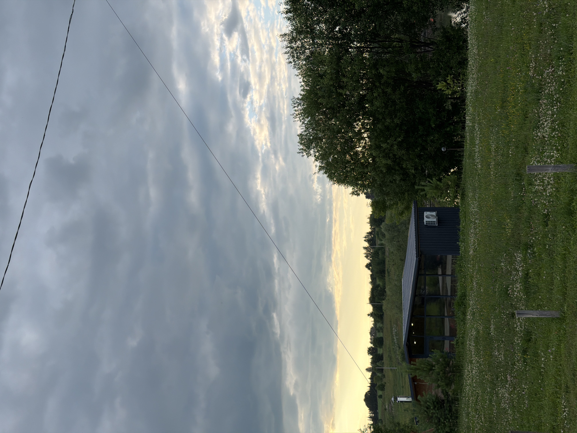 The image captures a rural landscape in Bol'shaya Ven'ya, Russia. The scene is dominated by a dramatic sky filled with a mix of dark, stormy clouds and lighter, brighter patches indicating sunset or sunrise. A single, thin power line stretches diagonally across the sky, adding a man-made element to the natural scenery.
In the middle ground, a modern-looking dark structure with large glass windows and an outdoor seating area is visible. This building appears to be a shed or a small cafe. An air conditioning unit is mounted on the roof of the structure. To the right of the building, a grassy field is dotted with small white flowers and is bordered by a wooden post.
The background features a dense line of trees, suggesting a wooded area or forest. The overall impression is one of a serene, yet potentially dynamic, natural environment at dusk or dawn. The lighting suggests it's either early morning or late evening. There are no people visible in the image, and no discernible text is present.