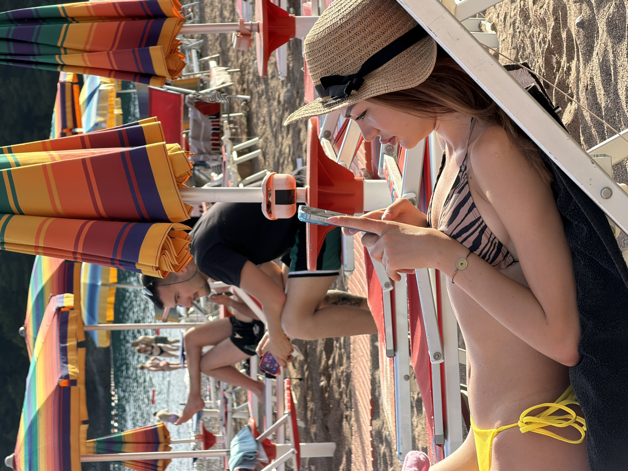 The image depicts a vibrant late-afternoon beach scene in Boreti, Montenegro. In the immediate foreground, a young woman with auburn hair, wearing a straw hat with a black ribbon, a striped bikini top, and yellow bikini bottoms, reclines on a red and white striped lounge chair. She is engrossed in a light blue smartphone held in her hands. A circular metallic charm bracelet is visible on her left wrist, and a black towel is placed beneath her on the chair.
Further back, towards the center-left, a man with a beard and dark hair sits on a similar red and white lounge chair. He is dressed in a black t-shirt and green and black shorts, holding a red smartphone. Sunglasses rest on his head, and his legs are crossed as he relaxes.
The background is dominated by numerous large, colorful striped beach umbrellas in various patterns, including combinations of orange, red, purple, yellow, green, and blue stripes. Beyond the umbrellas, the sea extends, dotted with several individuals swimming or wading, with two figures appearing to embrace in the water. The distant shoreline or hills are visible as a dark, indistinct silhouette. The sandy beach shows ripple patterns, and additional white lounge chairs are scattered across the scene. The lighting is warm and golden, characteristic of late afternoon sun, casting noticeable shadows.