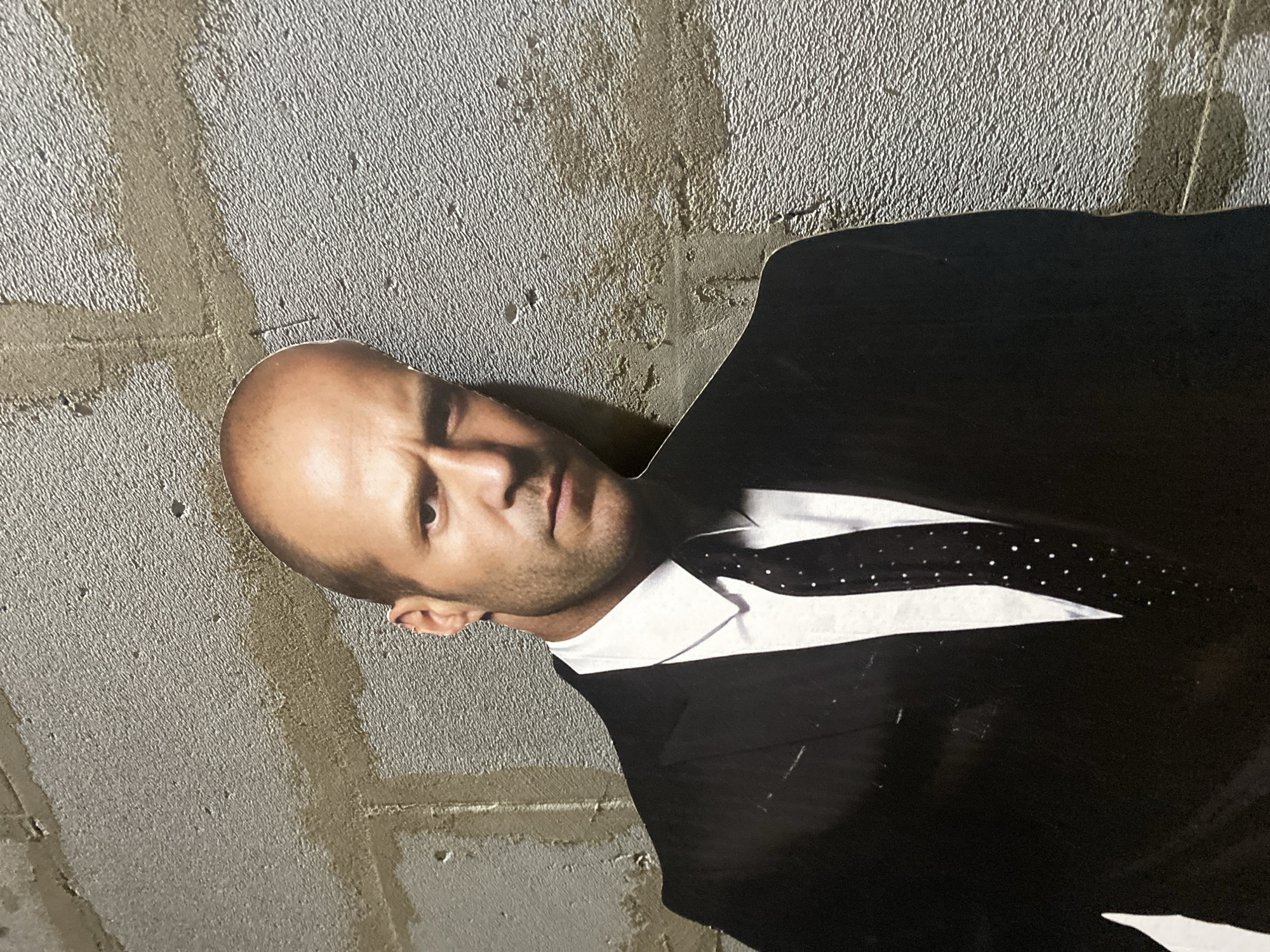 A portrait of a bald man in a suit and tie is adhered to an unfinished grey wall. The background wall appears to be textured and unpainted. The image is angled, with the top of the man's head pointing towards the top left of the frame. The portrait appears to be a cutout or poster.
There is no information given to suggest this image is located in the city of Chaikovsky, Russia.