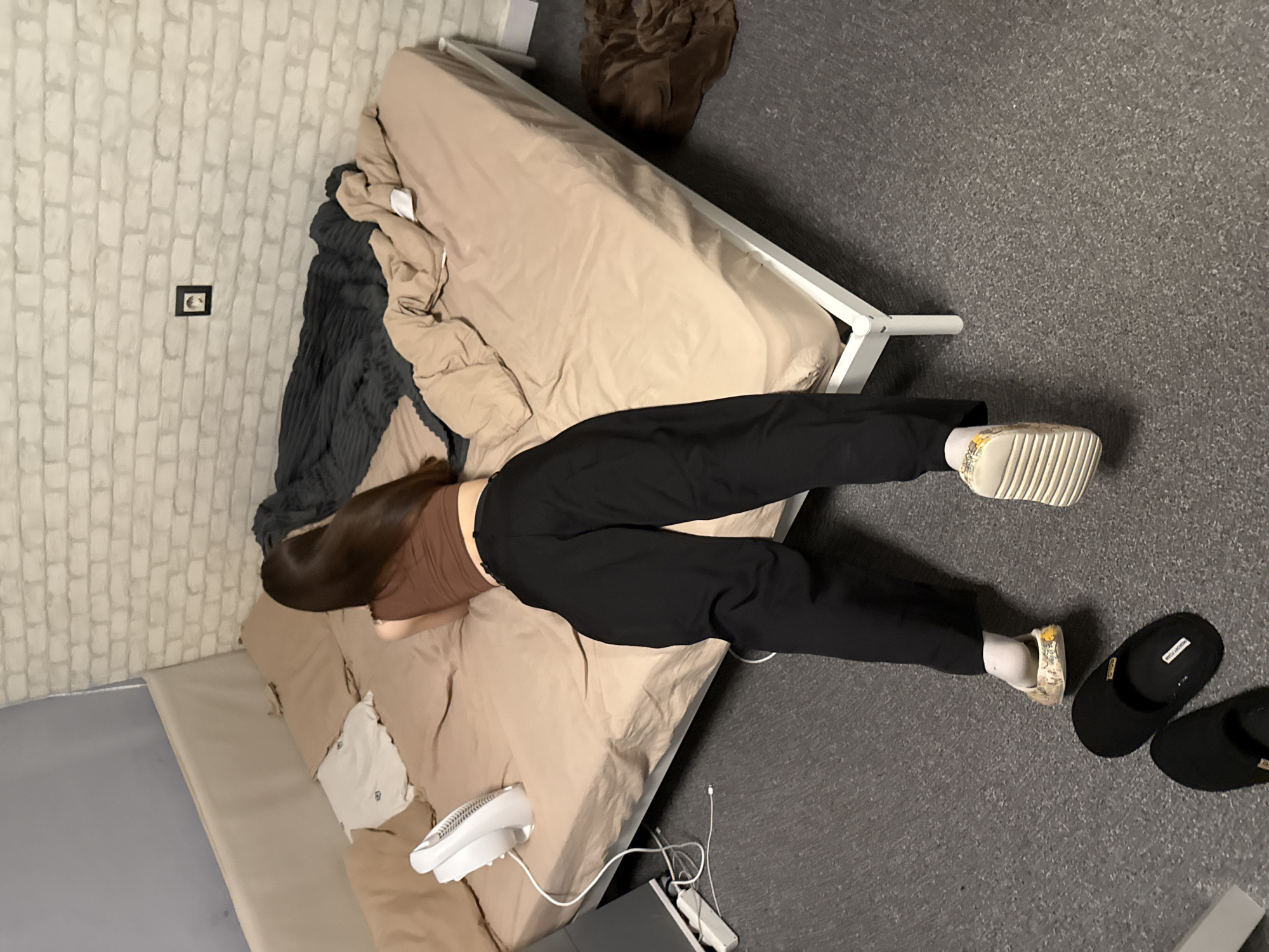 The image, taken in Sofia, Bulgaria, shows a person lying face down on a bed in an indoor setting. The person, with long dark hair, is wearing a brown top and black pants, white socks, and light-colored sneakers with patterned details. Their face is obscured by the pose.
The bed has a simple white frame and is covered with a beige fitted sheet. Some rumpled bedding, including a dark gray blanket and more beige fabric, is visible at the head of the bed. A small white fan and a charging cable are on the bed near the person's torso.
The floor is covered with a dark gray carpet or rug. Next to the person's feet, a pair of black slippers is visible, with the text "MEMORY FOAM" legible on one. On the floor beside the bed, a power strip and additional charging cables are present, along with what appears to be a laptop or tablet on the ground. A brown crumpled item is also on the floor near the head of the bed.
The wall behind the bed features white brick-patterned wallpaper. A small, dark-framed picture or artwork is mounted on the wall. Another bed or mattress with a light-colored surface is partially visible to the left of the main bed, positioned against the wall. The lighting suggests an indoor setting, likely with artificial light, without clear indications of the time of day or weather. The person appears to be resting or sleeping.