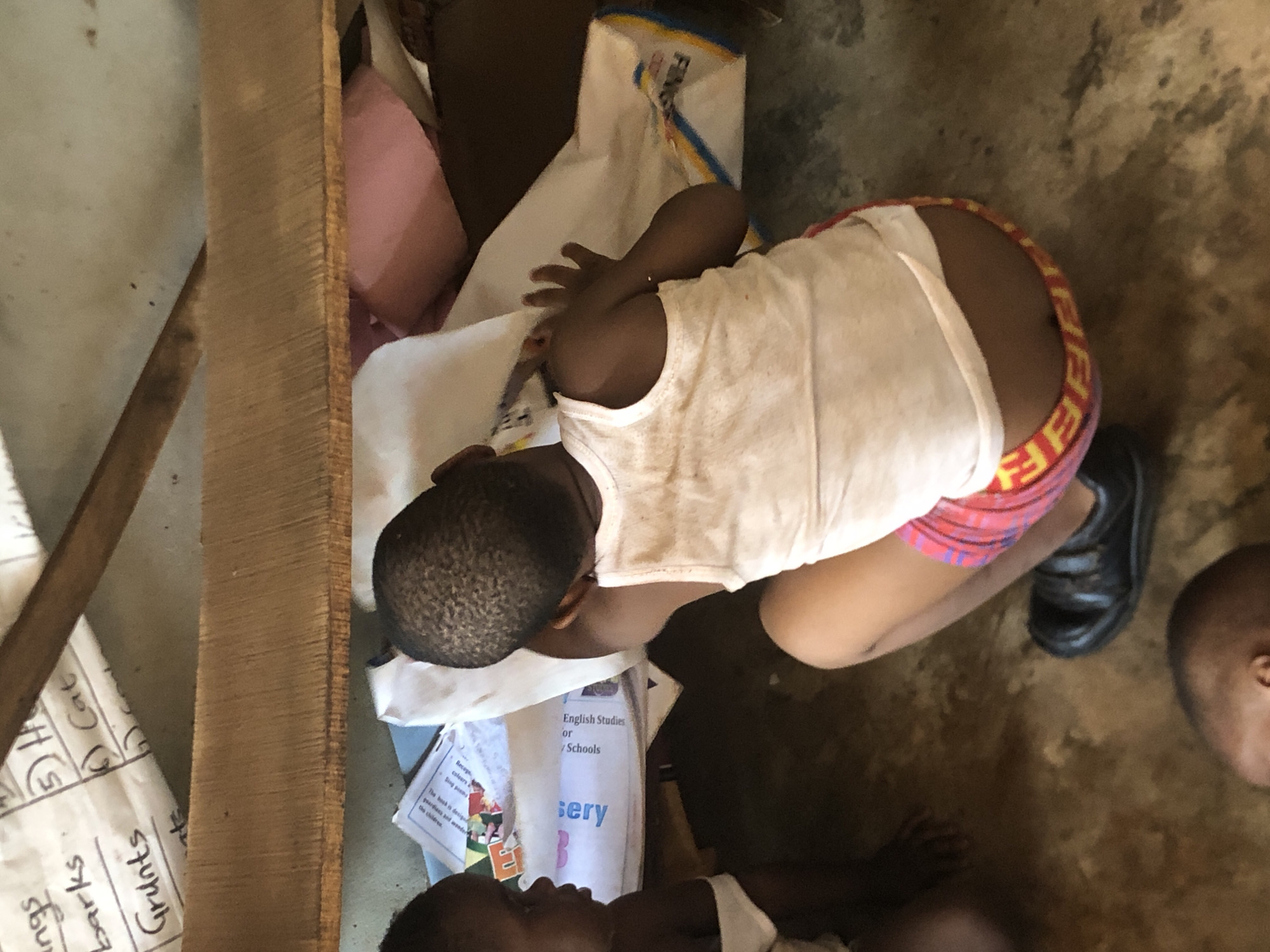 A top-down shot captures a scene within what appears to be a room in Ukwu-Abwa, Nigeria. The location is indicated by the presence of educational materials used in Nigerian schools. A child is kneeling, leaning over school books and papers, wearing a white undershirt and pink shorts with a distinctive, branded waistband. The child's hand rests protectively on the documents. A black shoe is visible near the child. Another child is lying down near the educational materials. Additional items, including a piece of lumber, a pink pillow, and a white bag, are also visible within the frame. The floor appears to be a textured, earthy tone.