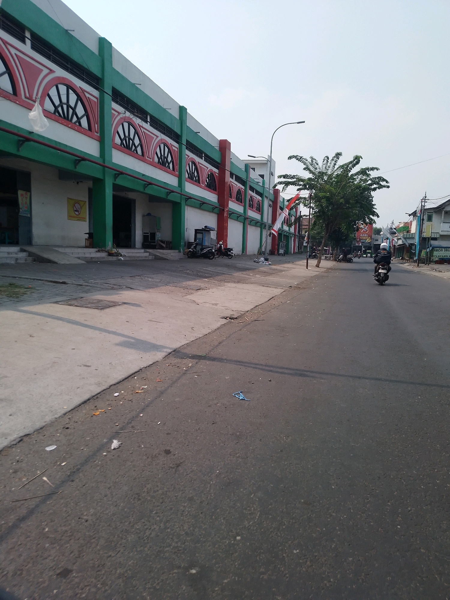 The image shows a street scene in Kertosono, Indonesia. On the left side of the street is a long, multi-story building with a green and white facade, adorned with red arched windows and green vertical pillars. A ramp leads up to the entrance of the building. Several motorcycles are parked in front of the building. A street lamp is visible above the building.
The street itself is paved with asphalt and appears to be a relatively busy thoroughfare. There are several vehicles on the road, including a person riding a motorcycle from right to left in the foreground. Trees and other buildings are visible on the right side of the street, some with shop signs. The sky is overcast. Debris, such as plastic bags and leaves, is scattered along the edge of the road and sidewalk. The overall scene depicts a typical streetscape in a town or city, with commerce and transportation evident.
