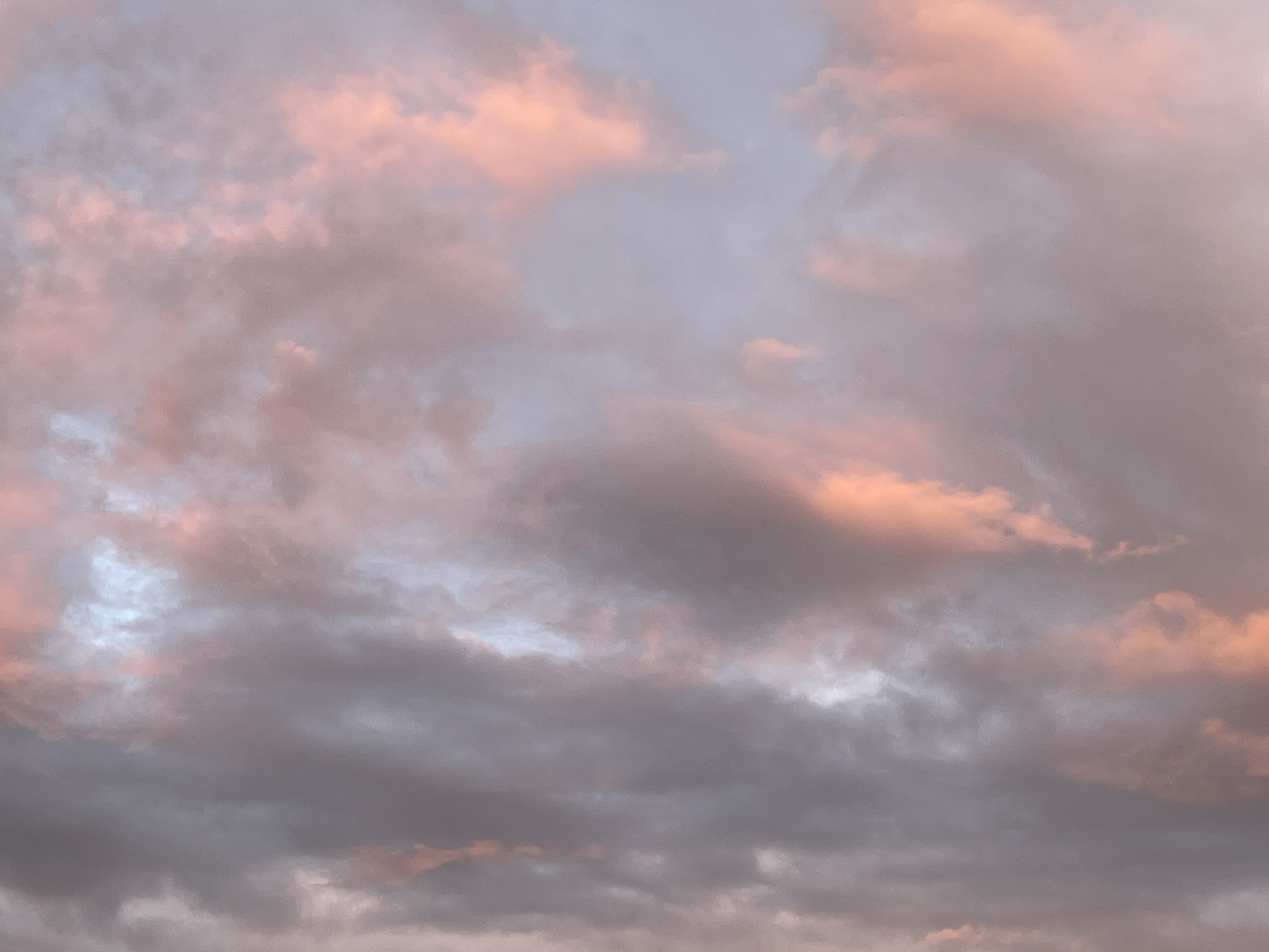 The image is a close-up, full-frame photograph of a cloudy sky at dusk, illuminated by soft, warm light. The clouds are a mix of pastel pinks, peaches, and grays, with hints of pale blue peeking through in some areas. The lighting suggests a sunset or sunrise. The texture of the clouds is varied, with some appearing wispy and translucent, while others are more dense and textured. There are no people, man-made structures, or geographical landmarks visible, making it impossible to determine a specific location, including in the city of Vilnius, Lithuania. The scene is entirely natural, focusing solely on the atmospheric conditions of the sky.