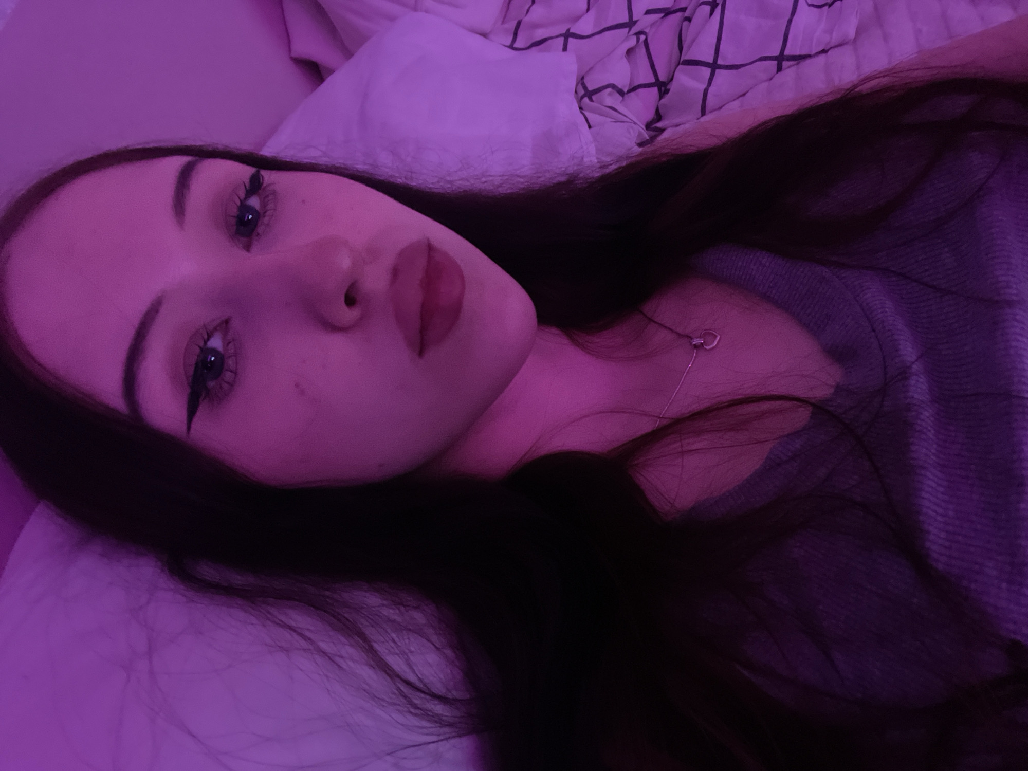 The image is a close-up selfie of a young woman with long, dark hair, lying on a bed. She is wearing a gray v-neck top and a silver necklace with a heart pendant. Her eyes are open and she has dramatic winged eyeliner. The lighting is predominantly purple, casting a shadow over much of the scene. The background shows a pillow and a duvet cover with a black grid pattern. The location of Votkinsk, Russia is not discernible from the image itself.