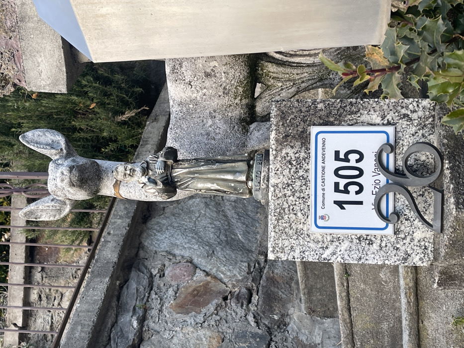 The image, oriented on its side, displays an outdoor scene featuring decorative statues and a house number plaque, set against a backdrop of stone steps and foliage, likely in Castione Andevenno, Italy.
In the center-left, a grey, stone statue of a donkey or mule is prominent. Beside it, or slightly in front, stands a smaller, silver-colored statue of a robed figure, identifiable as "BEATO PIO" (Blessed Pio) from the text on its base. This figure appears to be holding a small object.
To the right of the statues, a white rectangular house number plate with a blue border is affixed to a textured stone block. The plate clearly reads: "Comune di CASTIONE ANDEVENNO", the number "1505", and "Via Ezio Varani". A small crest or logo is also visible on the plate. Below this plate, separate dark grey metal numbers "26" are mounted on the stone surface.
The background shows grey stone steps and a dark metal railing to the left, suggesting a pathway. Various green bushes and plants, including holly, are visible around the statues and stone structures. The scene is illuminated by daylight, casting distinct shadows, indicating a clear, sunny day. The overall setting combines rustic stone elements with religious iconography and clear residential identification markers.