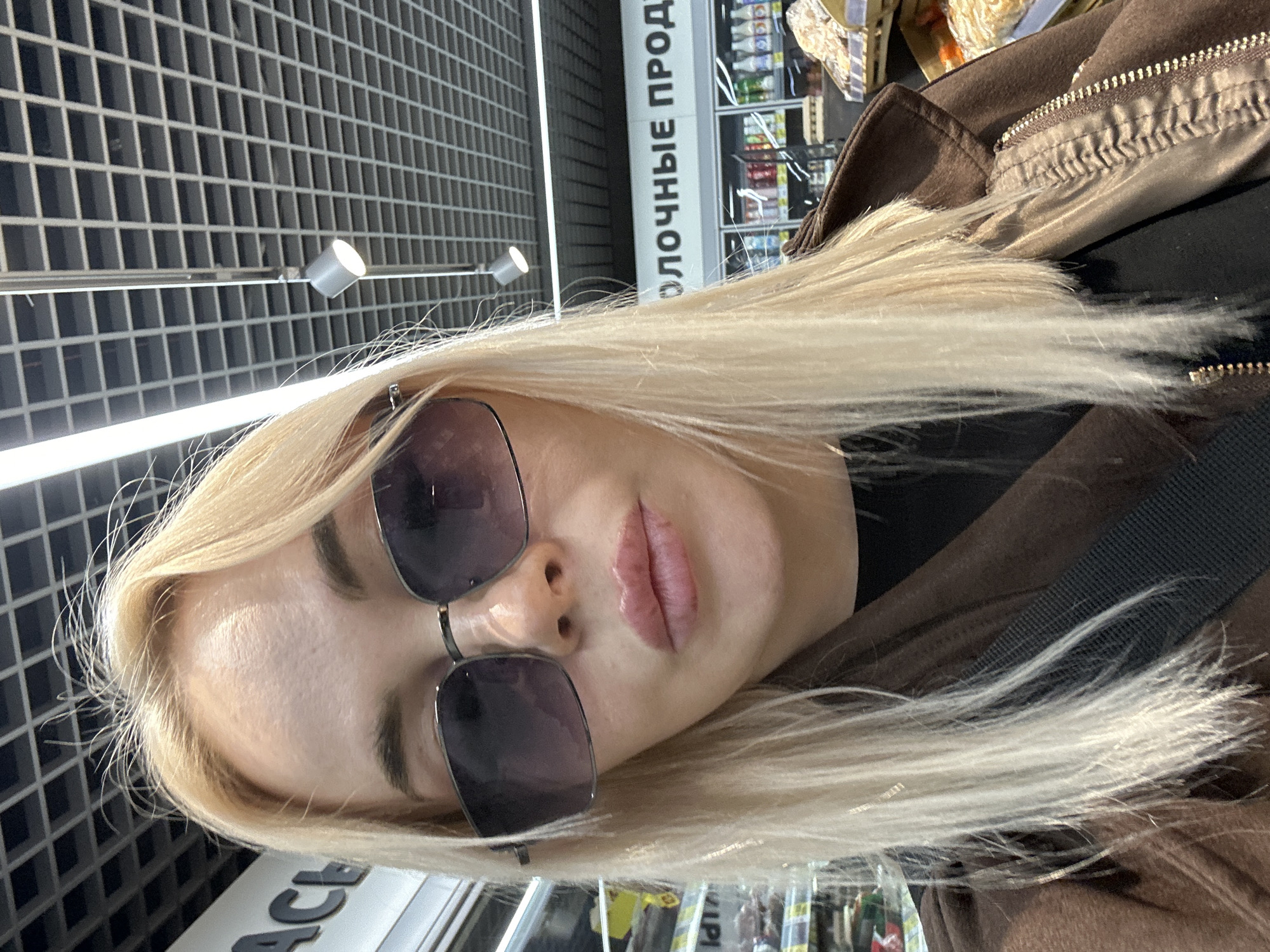 A woman with blonde hair and sunglasses is in a supermarket in Megion, Russia. She is wearing a brown jacket. The supermarket has shelves of products and a grid ceiling with lights. The sign above the shelves is written in Russian.