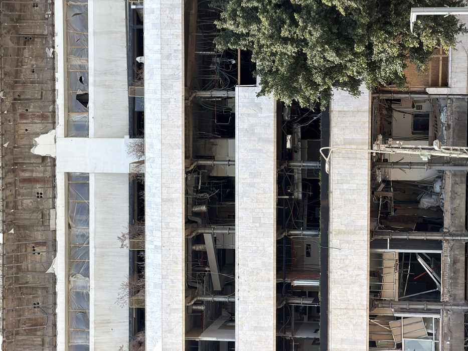 The image displays a multi-story building in Tel-Aviv, Israel, undergoing deconstruction or in a severe state of disrepair. The uppermost visible section features exposed concrete and rebar, indicating heavy structural dismantling or damage. Below this, multiple floors display a dirty white facade, with windows either shattered, missing, or covered by plastic sheeting. Patches of dry, sparse vegetation are present on several external ledges. The intermediate and lower levels are gutted, exposing structural columns, beams, extensive tangled electrical conduits, and ventilation ductwork. Construction debris, including fractured glass and discarded materials, is scattered throughout the visible interior spaces. A large green tree partially obscures the right side of the structure. A street light pole, equipped with a single lamp, is positioned in the lower-middle foreground. No persons are visible within the frame.