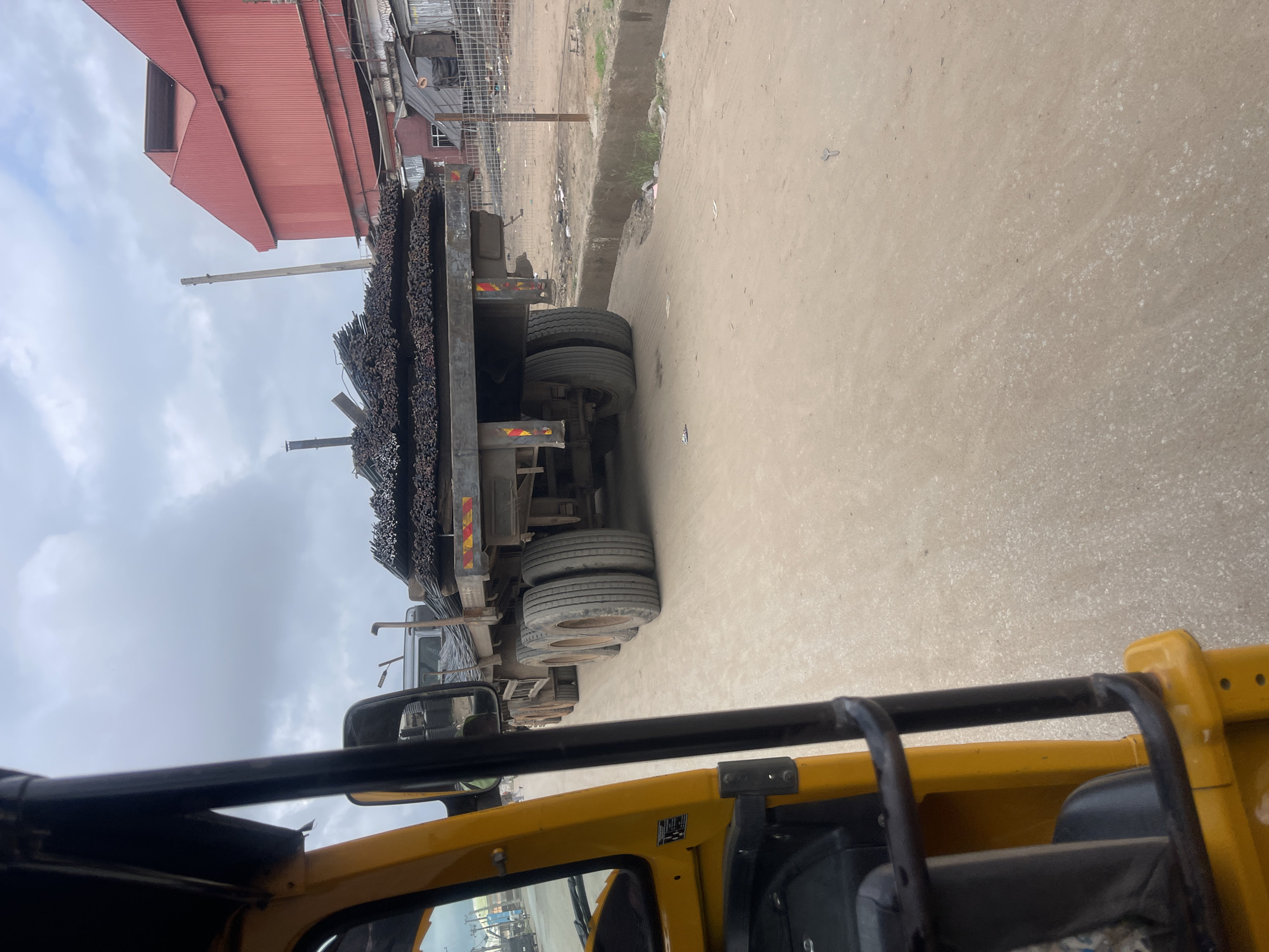 A flatbed truck loaded with rebar is parked on a dirt road in a commercial or industrial area in Idemili, Nigeria. The truck is seen from the perspective of a yellow auto-rickshaw. In the background, a large red industrial building with a corrugated metal facade is visible, along with a chain-link fence and some shacks. The sky is overcast. The road is dusty and appears to be under construction or poorly maintained. There are no people clearly visible in the image interacting with the truck or the surrounding environment.