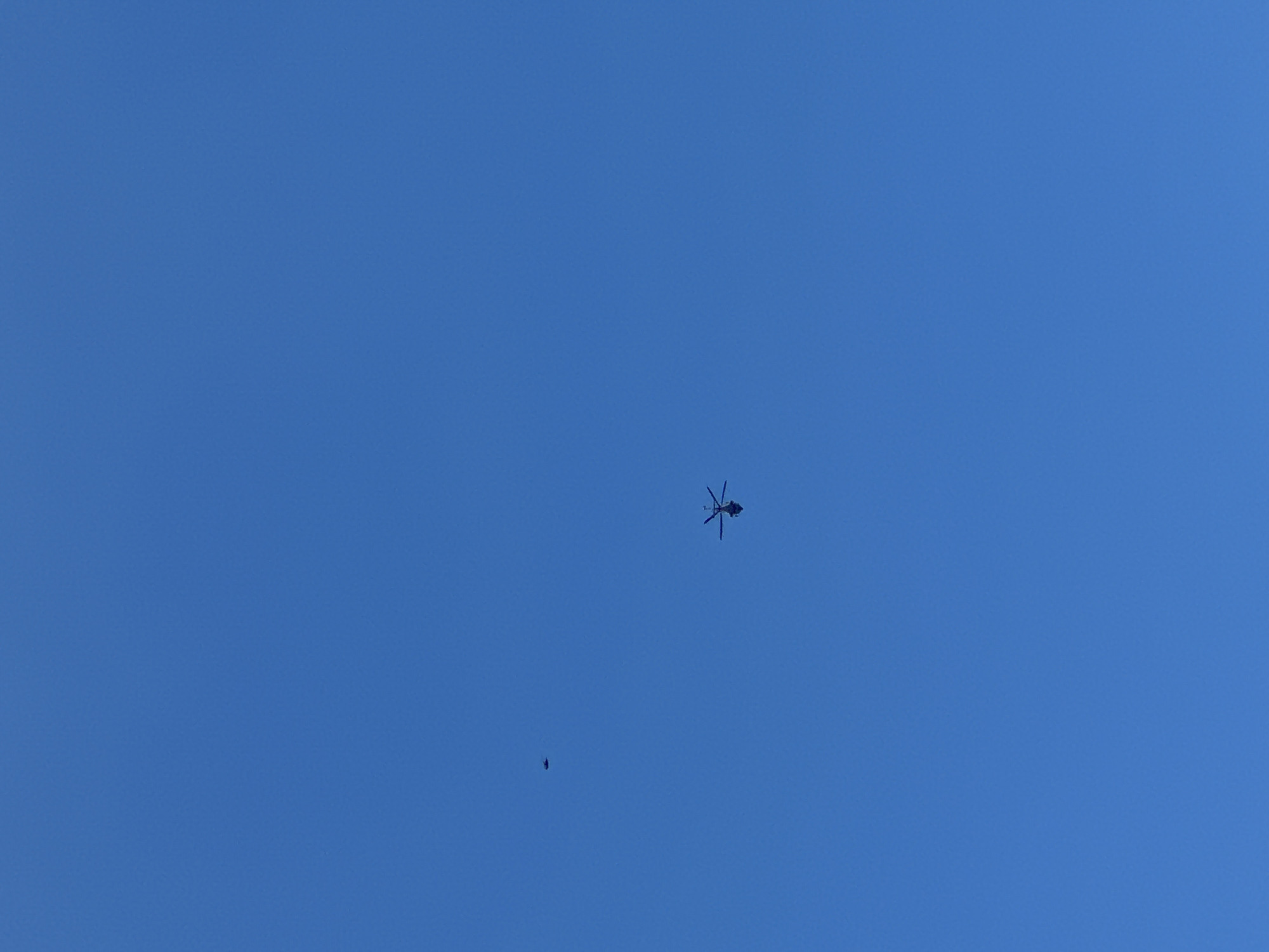 The image shows a clear, blue sky. In the center of the frame and slightly towards the top, there is a dark helicopter. Below and to the left of the helicopter, there is a smaller dark object. The location, Sant Feliu de Guíxols, Spain, is inferred due to the prompt request, however, there is no data visible in the image that confirms the location.